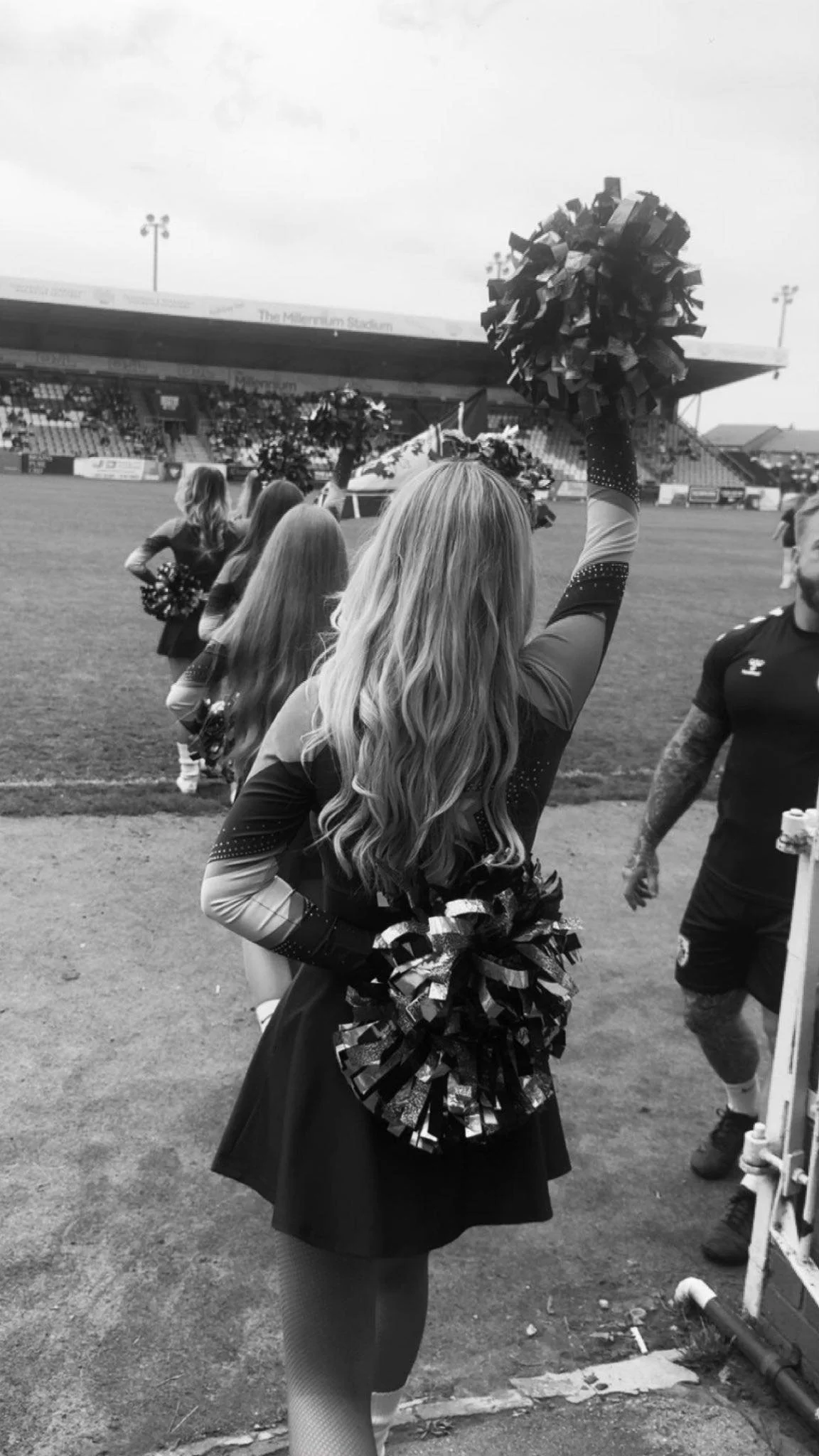 Cheerleader holding pom-poms at a sports stadium, seen from behind, with others in the background.
