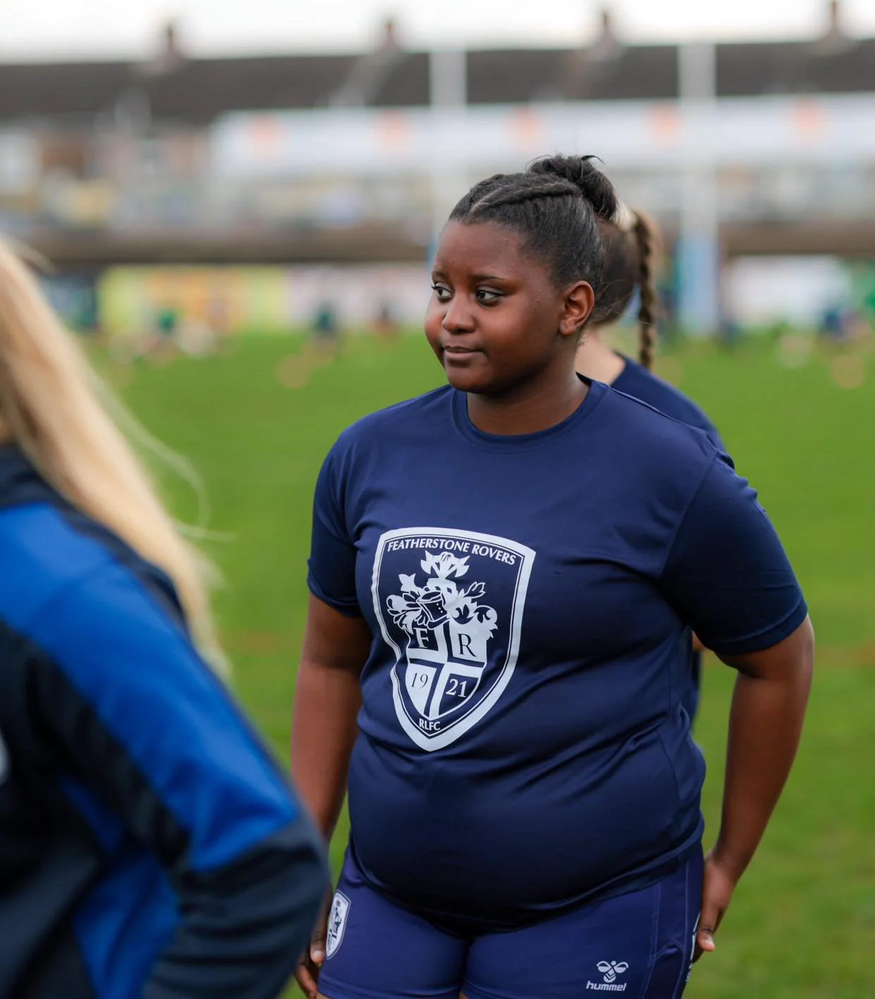 A female soccer player wearing a blue Featherstone Rovers jersey with a logo, standing on a grass field with a blurred stadium background, engaging in conversation with another person.