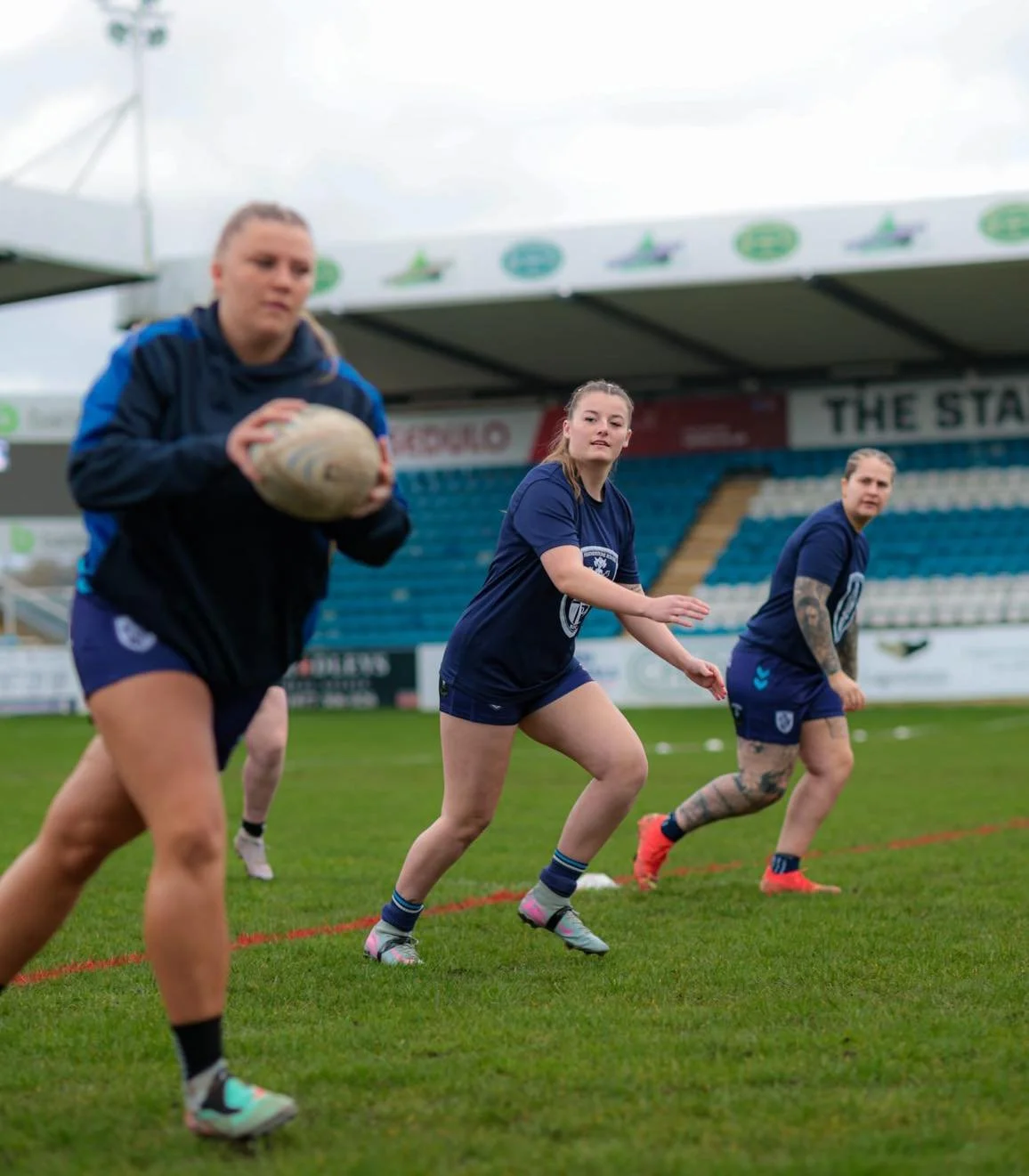 Women practicing rugby in a stadium, one holding a rugby ball, all wearing athletic gear.