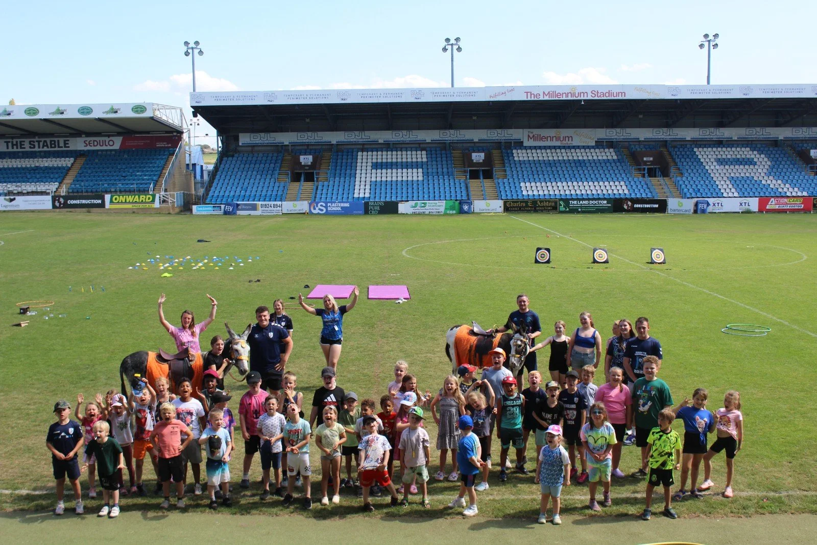 Group of children and adults on a sports field, some with ponies, in a stadium with seats and archery targets in the background.