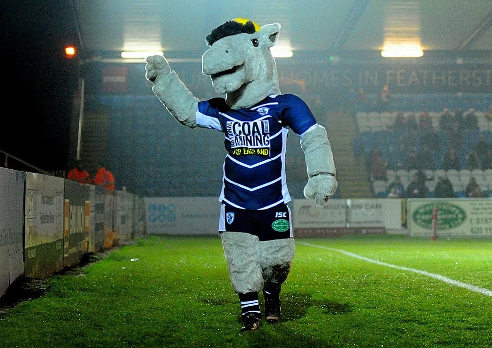A sports mascot dressed as a horse or unicorn in a blue and white rugby jersey, standing on a sports field. The mascot has a large head with a friendly face and black hair, and appears to be waving or pointing.