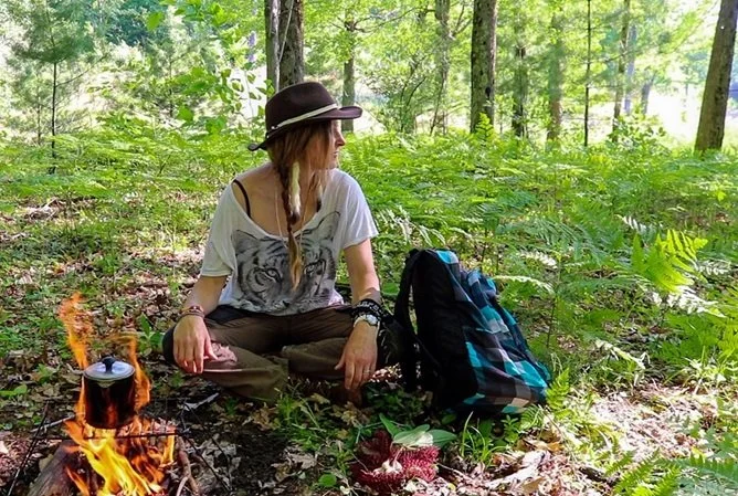 Young woman sitting on forest ground near a small campfire with a backpack, surrounded by trees and greenery.
