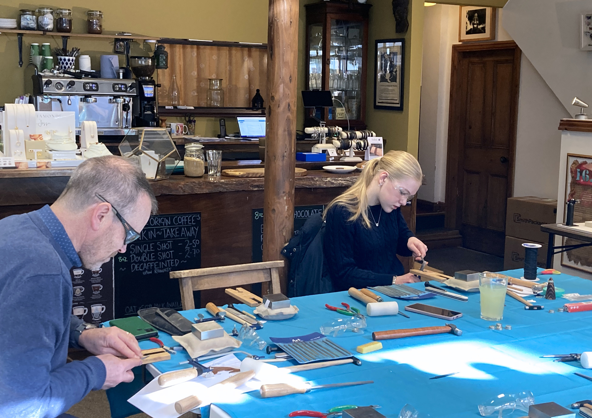 People making jewelry at a craft workshop with various tools and jewelry pieces on a blue tablecloth inside a cozy cafe or shop.