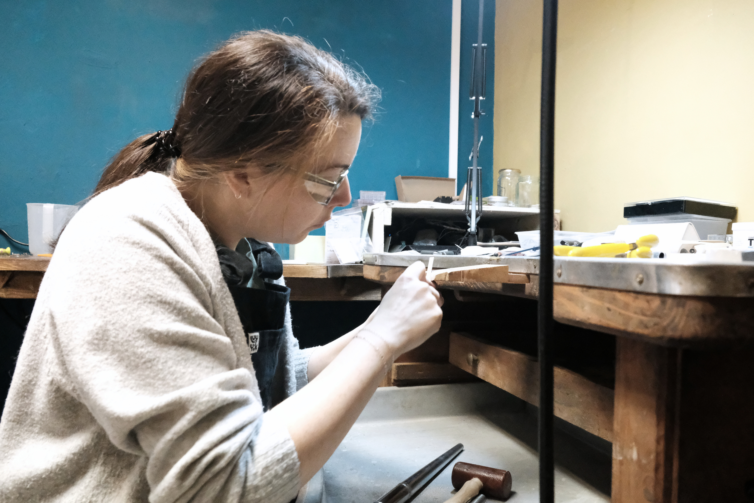 A woman with glasses working on a craft or project at a workbench in a workshop, with tools and supplies on the table.