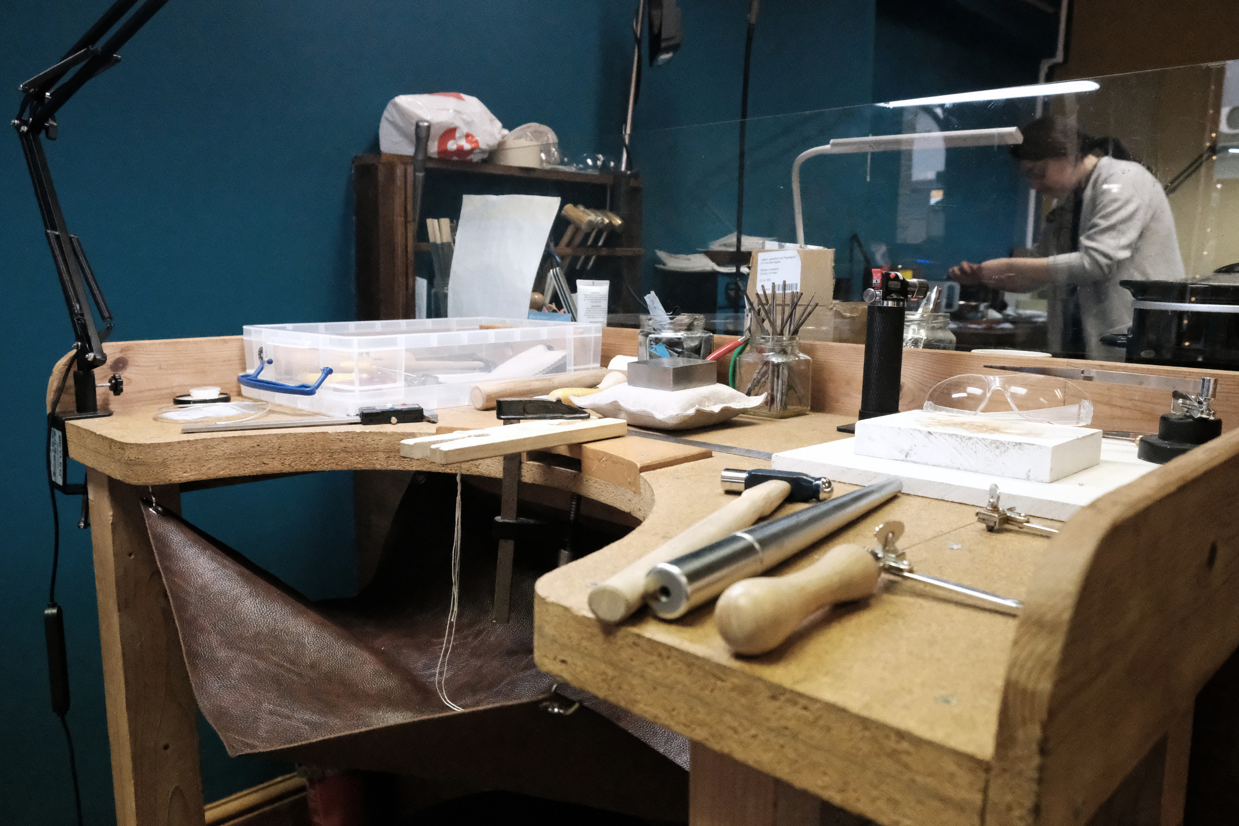 A woodworking workshop with a workbench holding various tools and supplies, separated from the worker's side by a glass barrier.
