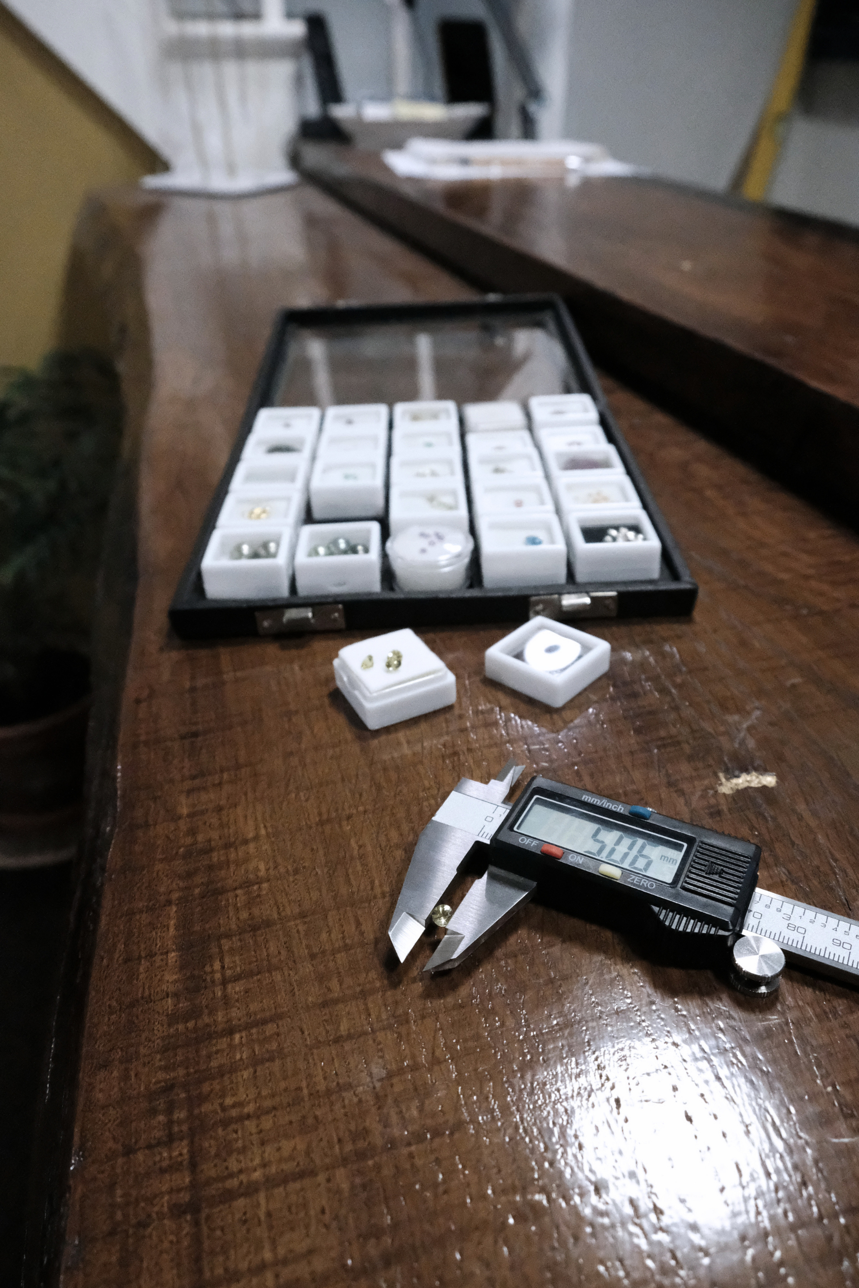 A wooden table with a black container holding small decorative objects and jewelry, along with a digital caliper measuring an item and other containers with jewelry pieces in the background.
