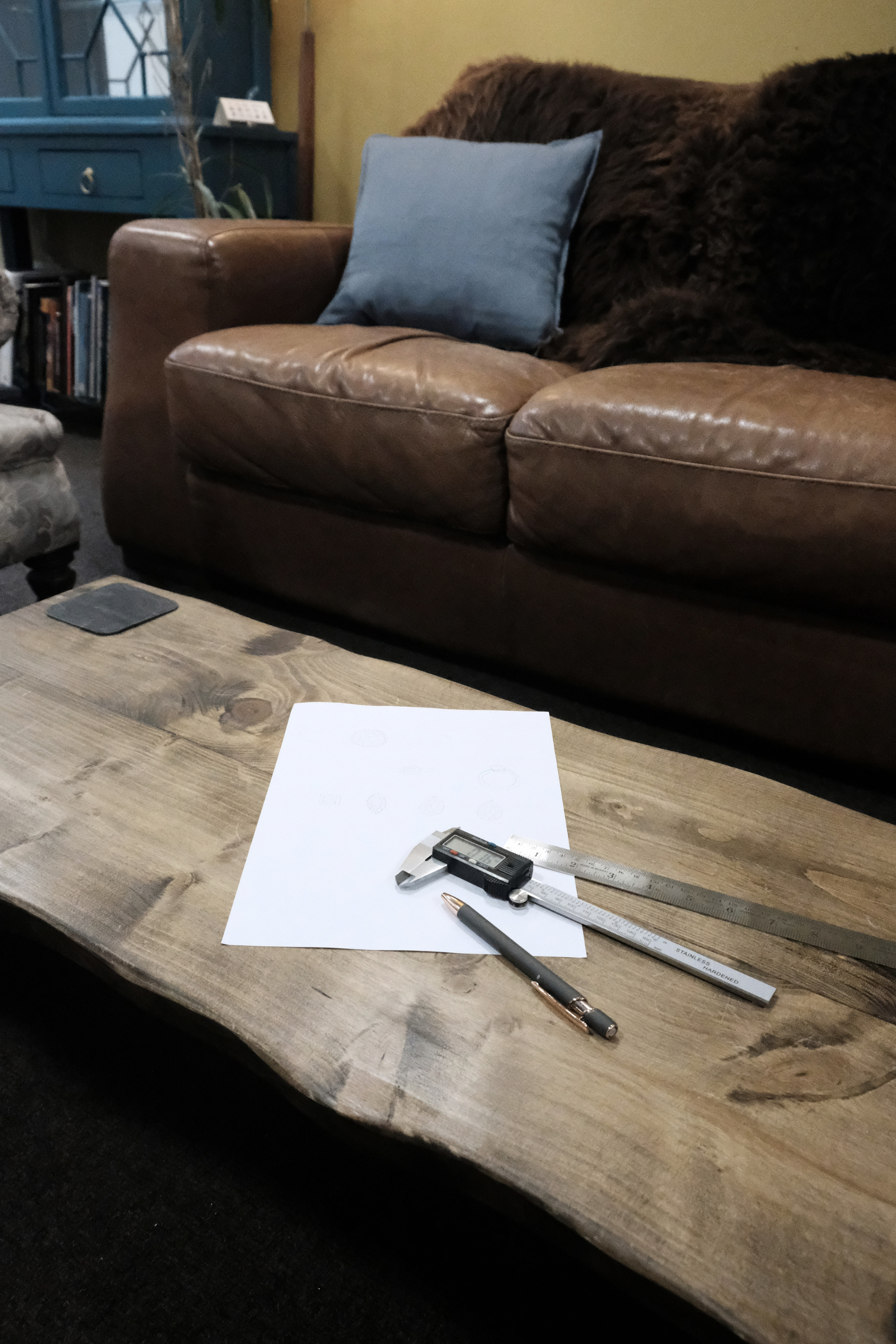 A wooden table with a piece of white paper, a digital caliper, a pen, and a ruler on it. In the background, there is a brown leather sofa with a blue cushion and a dark furry throw, along with a dark-colored shelf and a chair.