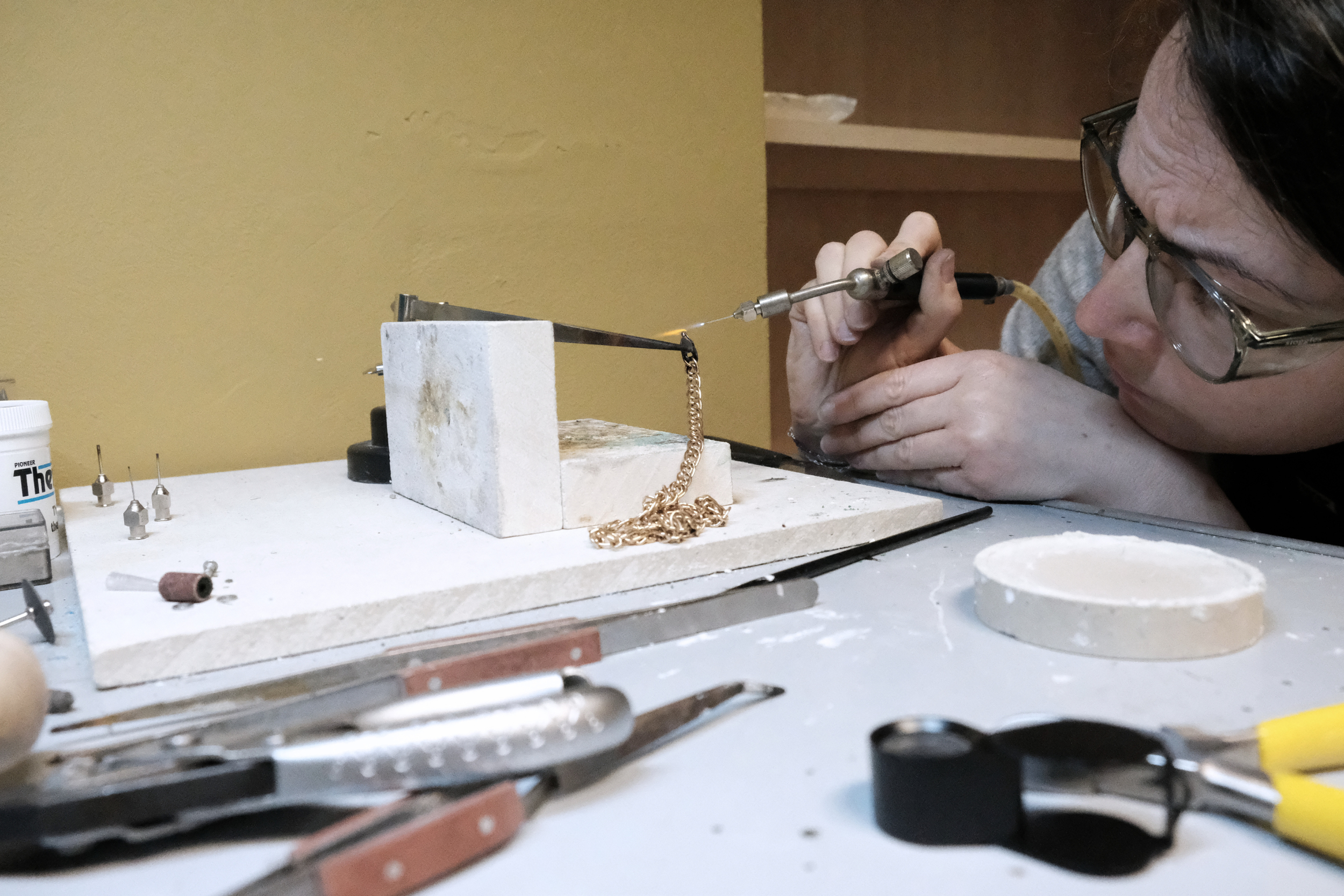 A person working on jewelry or metalwork, using a precision tool on a small chain, with various jewelry tools and materials on the work surface.