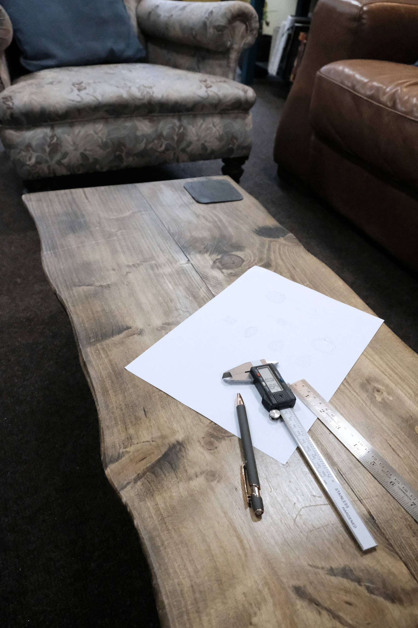 A wooden table with drafting tools, including a ruler, pen, and digital caliper, a sheet of paper with sketches, and a pen, with sofas in the background.