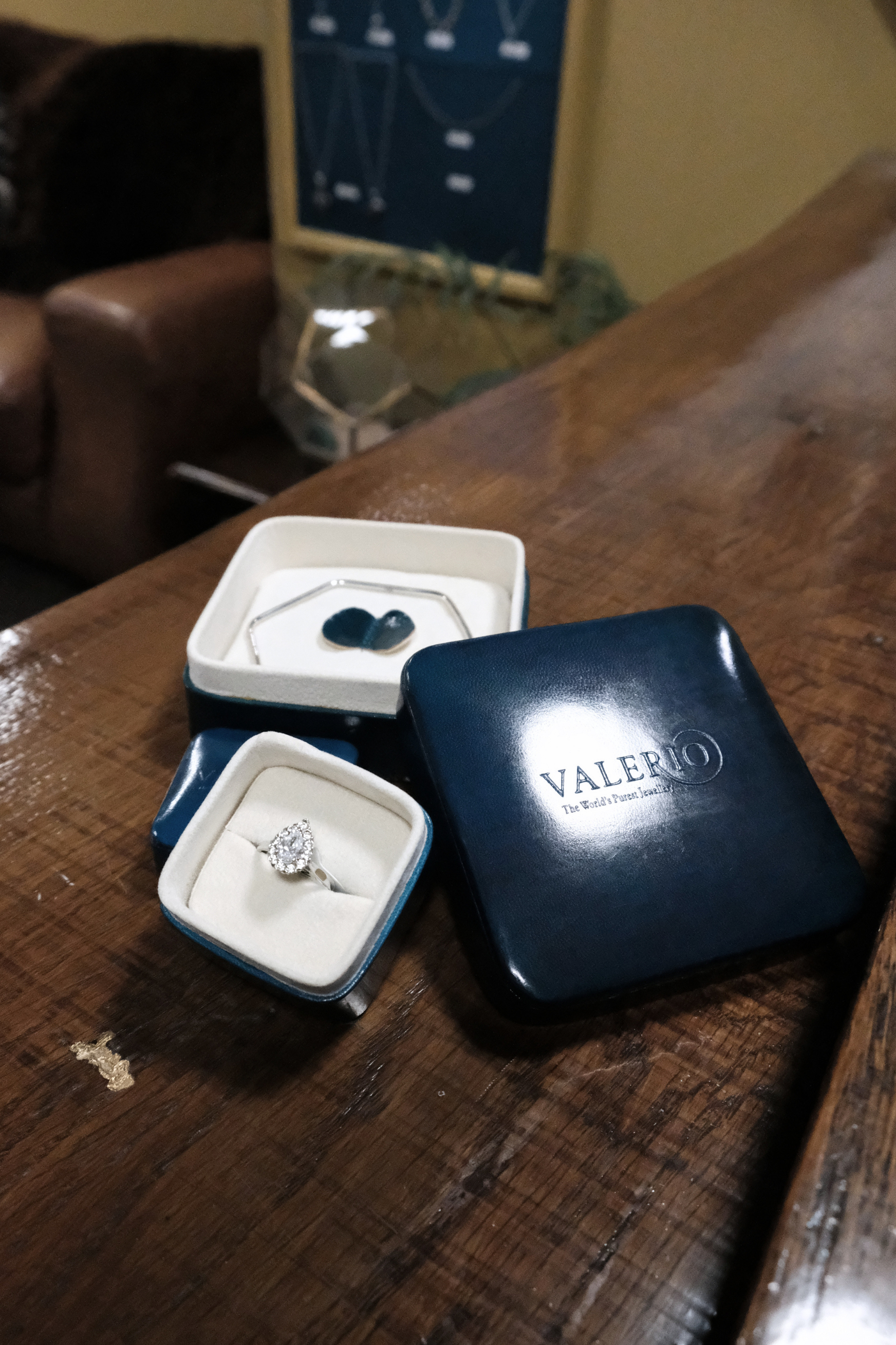 A wooden table with jewelry boxes, one containing a diamond ring, and a Valerio jewelry box. In the background, there is a brown leather chair and a blue corkboard on the wall.