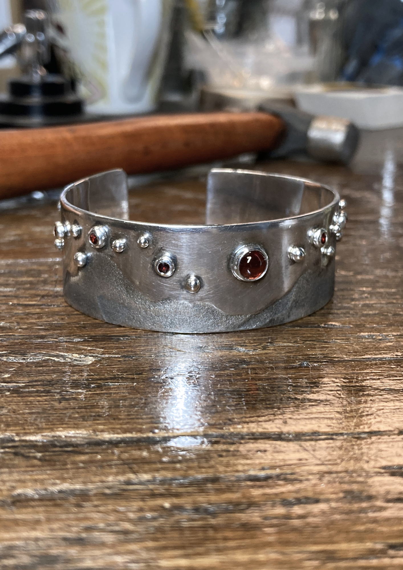 A wide silver cuff bracelet with embedded red gemstones resting on a wooden surface, with various kitchen or workshop tools blurry in the background.