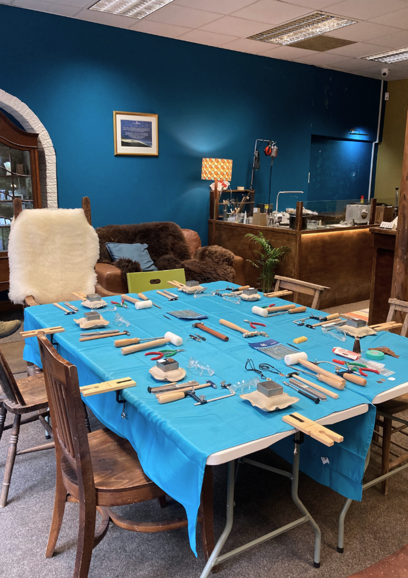 A table covered with blue cloth, filled with tools like hammers, pliers, and clamps, set in a room with a blue wall, a brown couch with a pillow, and a framed picture on the wall. The room appears to be a workshop or craft space.