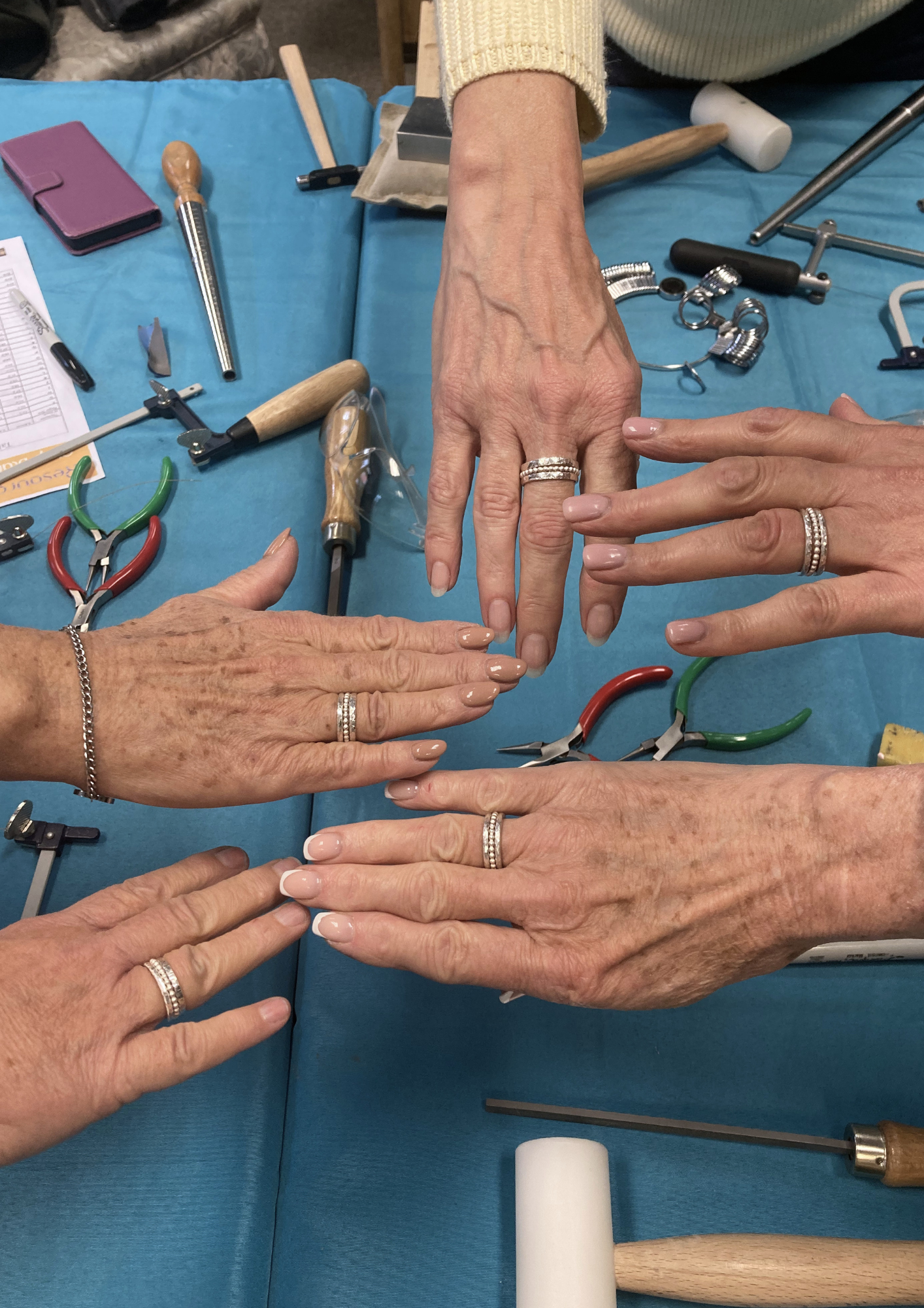 Multiple hands wearing rings are placed on a blue table with various jewelry-making tools and supplies, including pliers, hammers, and measuring tools.