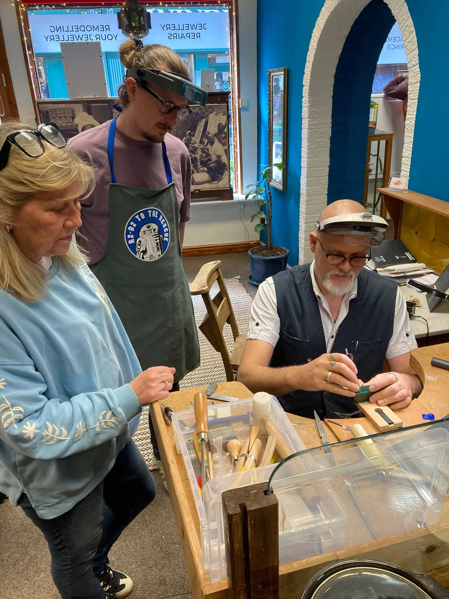 Three people working on woodworking projects in a workshop. One woman is standing and looking at her project, while a man is seated and carving or shaping wood. The third person is standing nearby, observing. The workspace has woodworking tools, a plastic container with wooden tools, and an indoor plant, with a colorful wall and a window in the background.