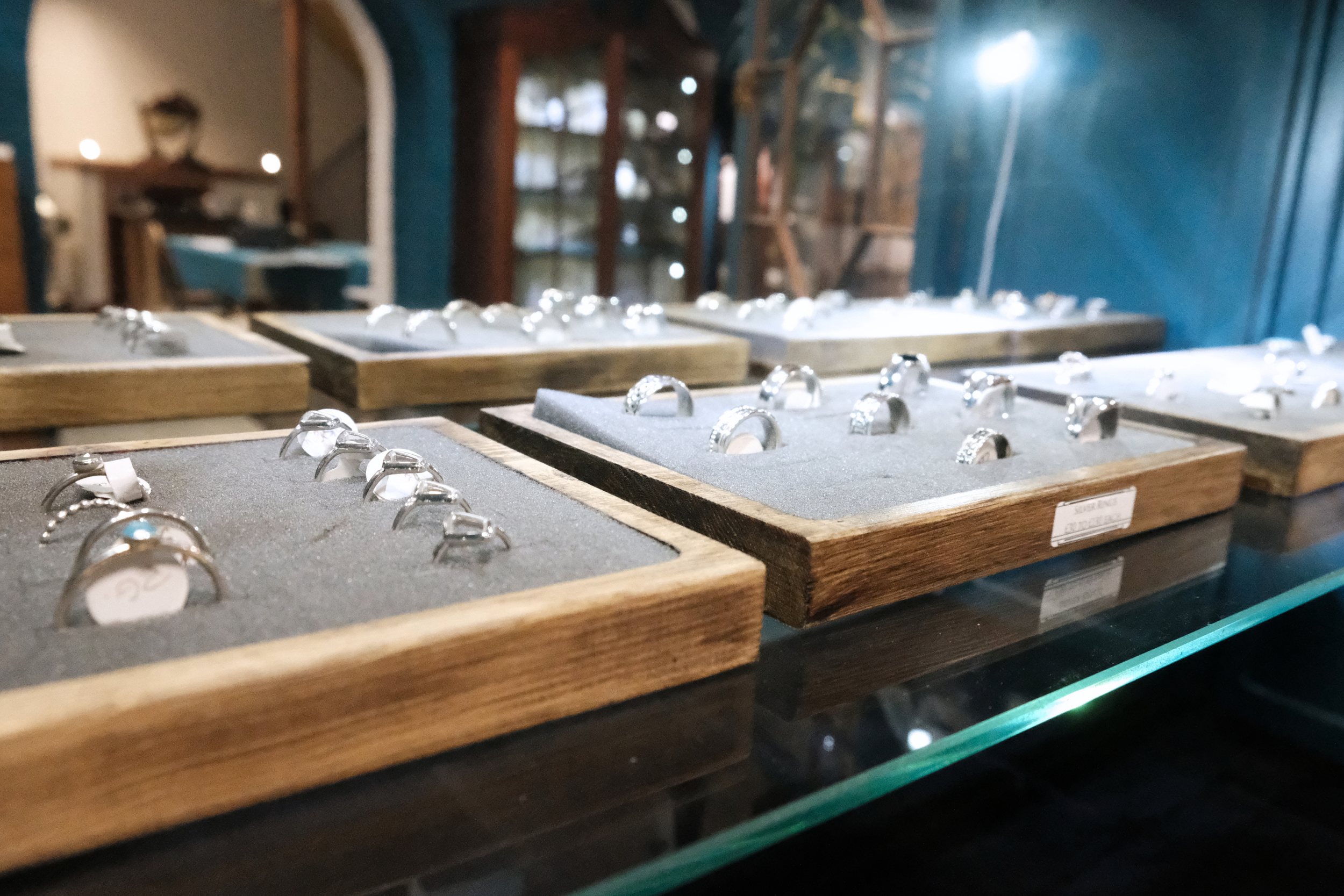 A display of various silver rings on wooden trays inside a jewelry store, with a blurred background and a blue wall.