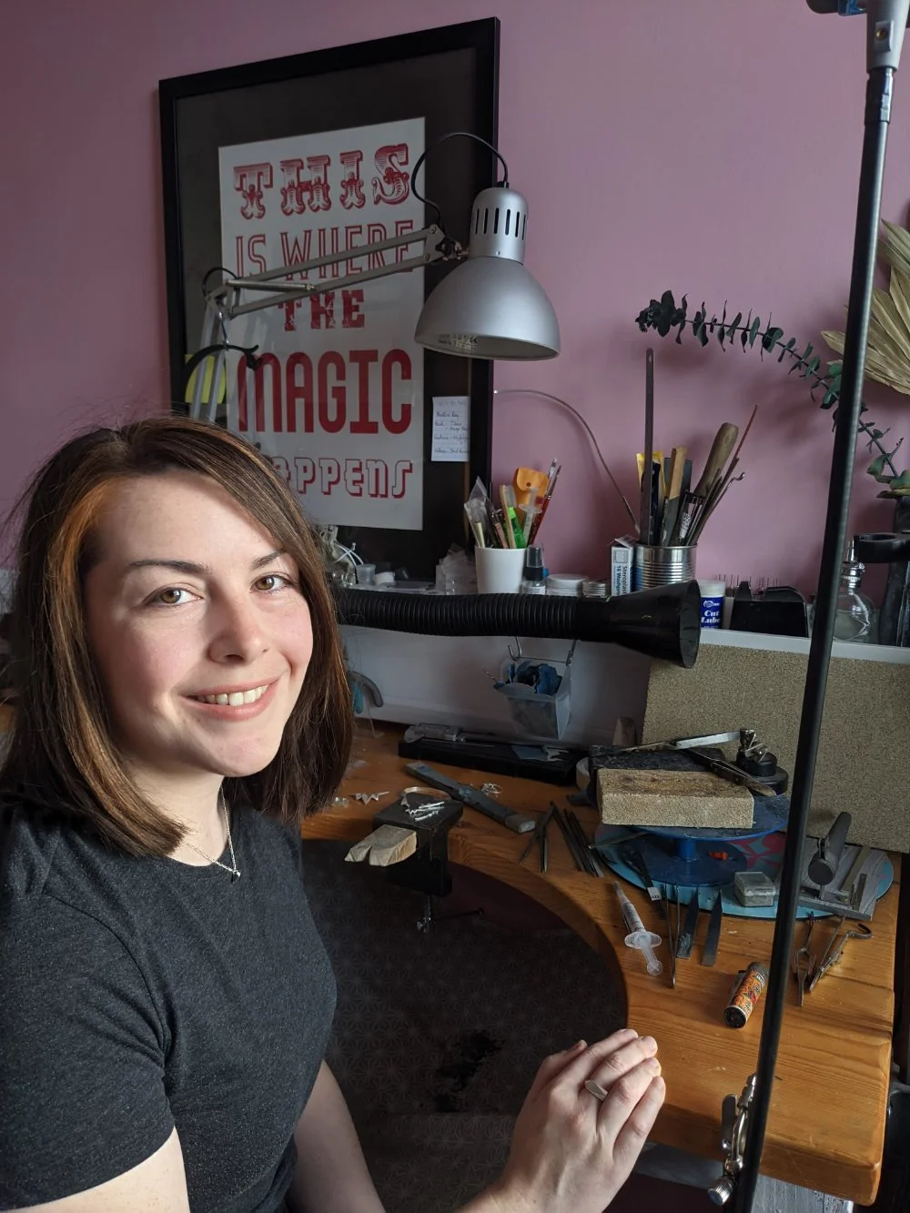 A woman sitting at a cluttered workbench in a jewelry making or crafting space, smiling at the camera with tools and supplies scattered around.