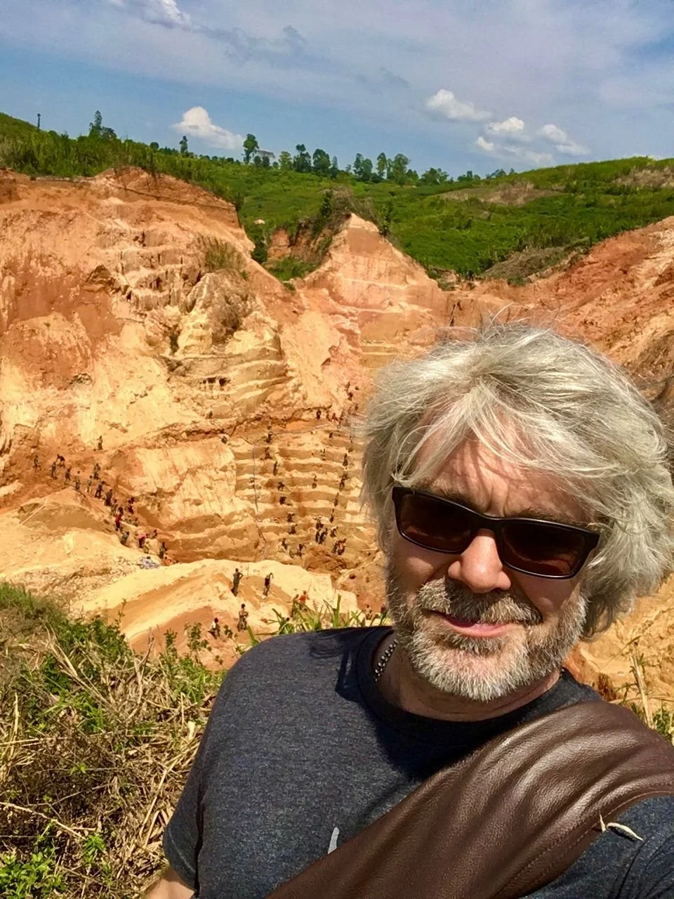 A man with gray hair, beard, dark sunglasses, wearing a dark t-shirt and a brown shoulder bag, taking a selfie in front of a colorful canyon with steep stairs and numerous tourists.