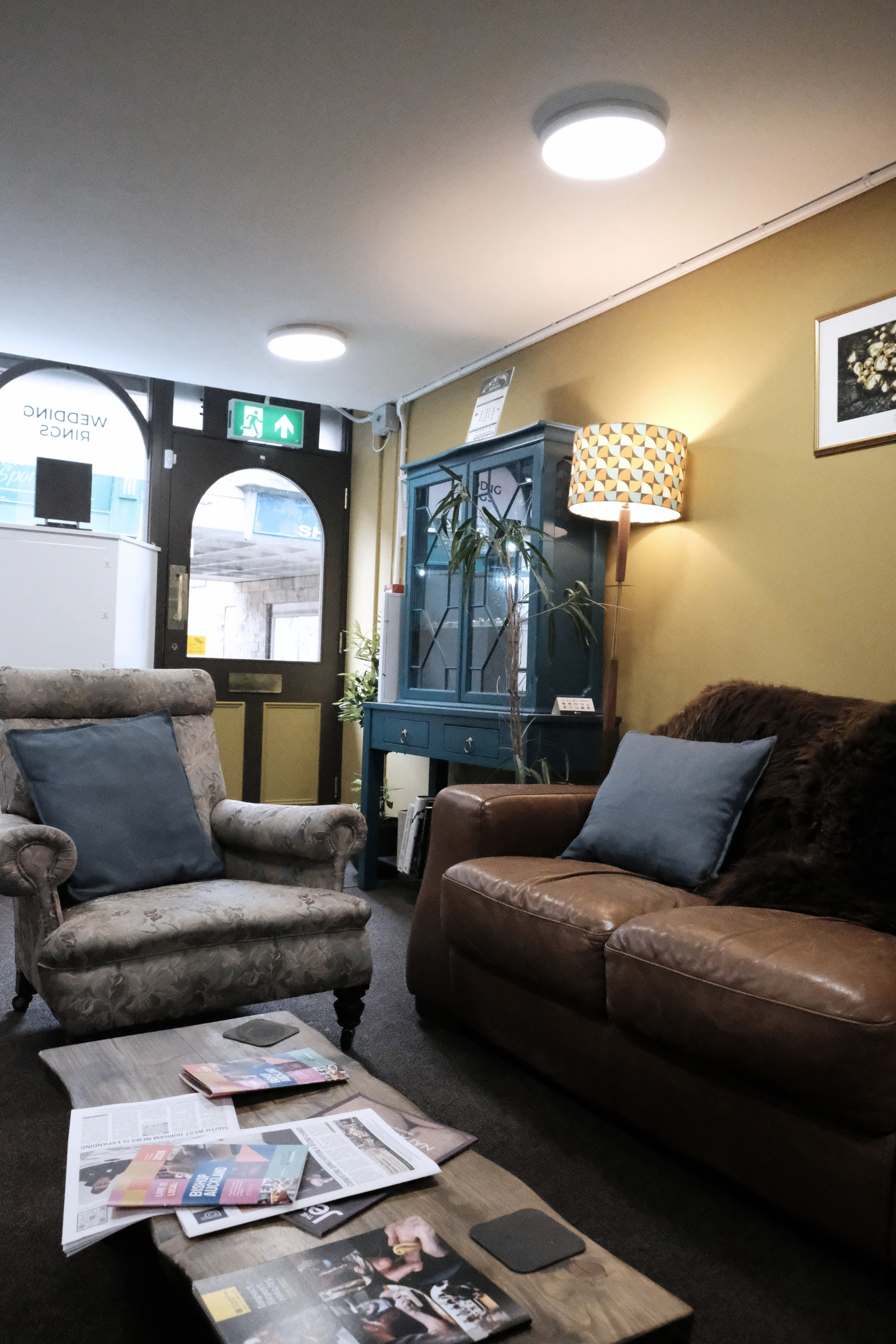 A cozy waiting room with vintage armchairs, a coffee table with magazines and newspapers, a blue cabinet with plants, and a green emergency exit sign above a door. Soft lighting and framed artwork on the wall create a warm atmosphere.