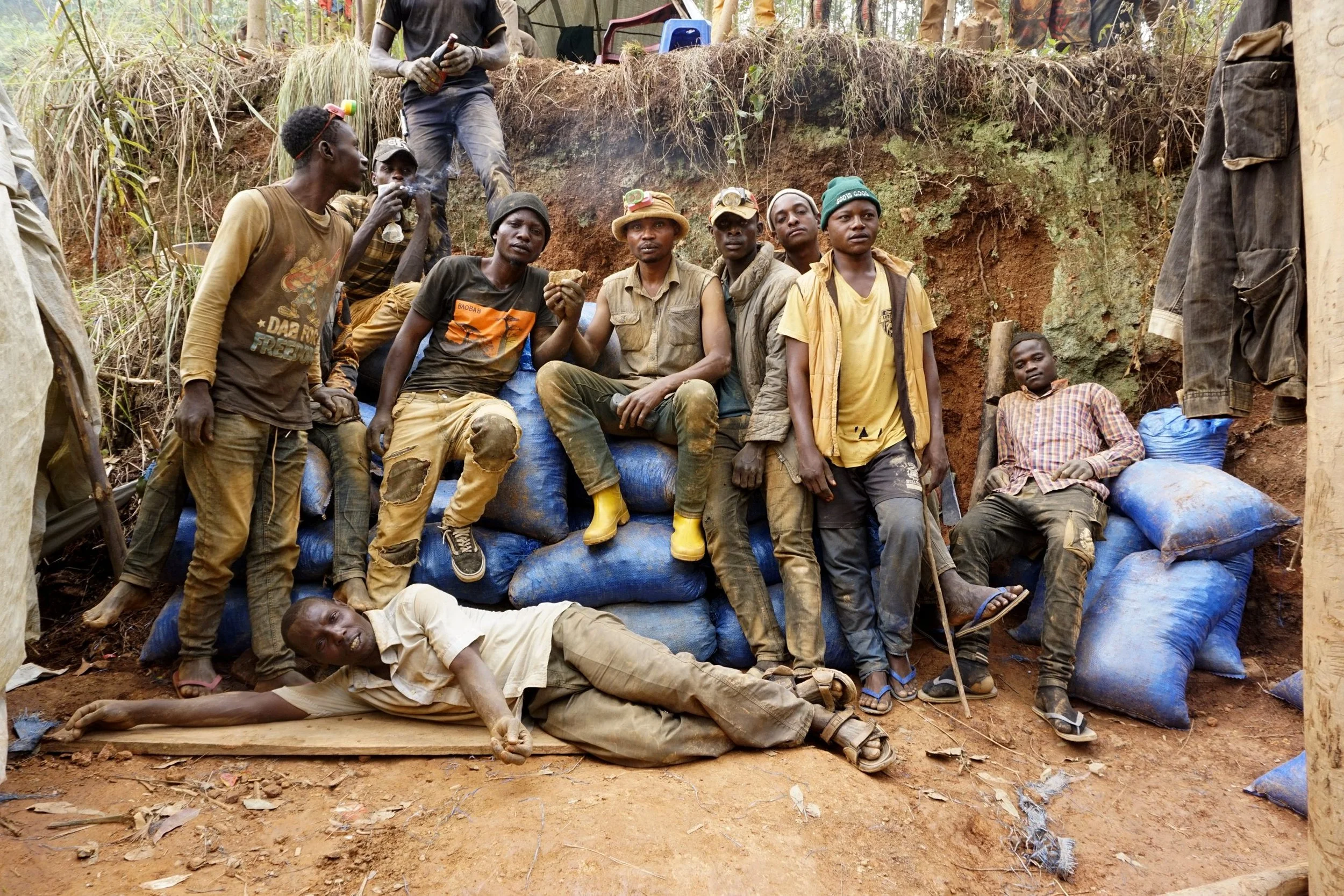 A group of people, mainly young men, gathered outdoors in a rural setting, sitting and lying on bags filled with earth or sand. They are dressed casually and appear to be resting or posing, with some holding bottles or food. The background shows a dirt hillside with sparse vegetation.
