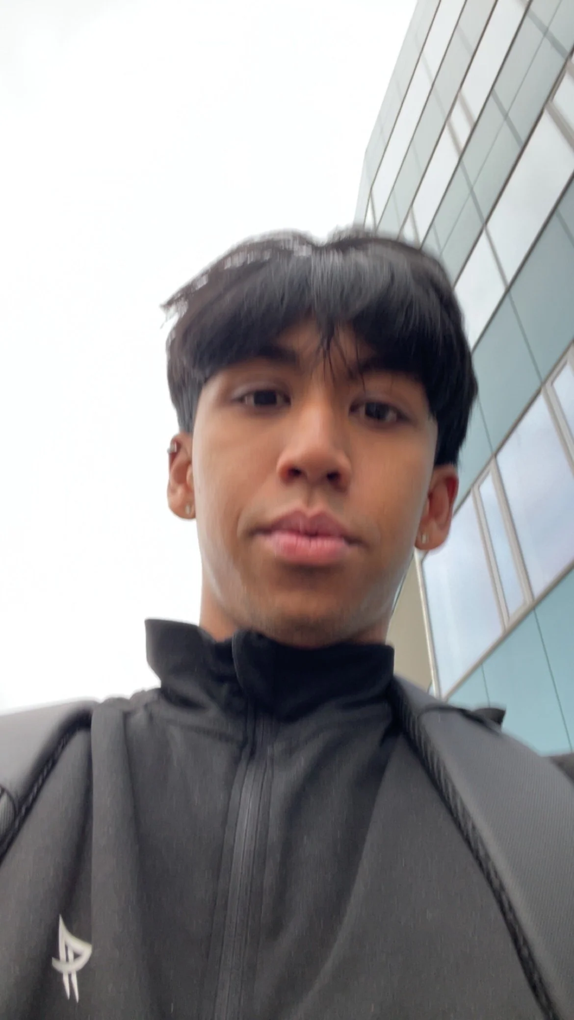 Young man with black hair and earrings taking a selfie outside a modern building with glass windows.