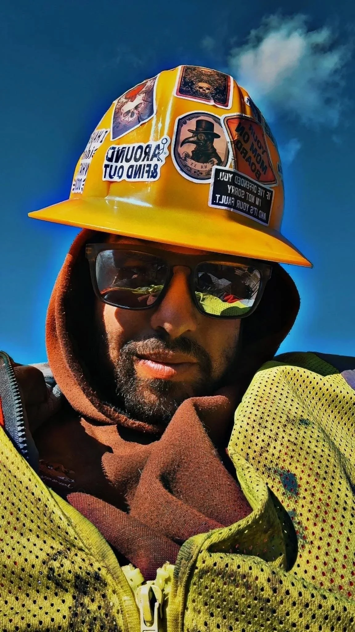 A person wearing a yellow safety helmet with various stickers, sunglasses, a brown hoodie, and a yellow safety vest, outdoors against a blue sky with some clouds.
