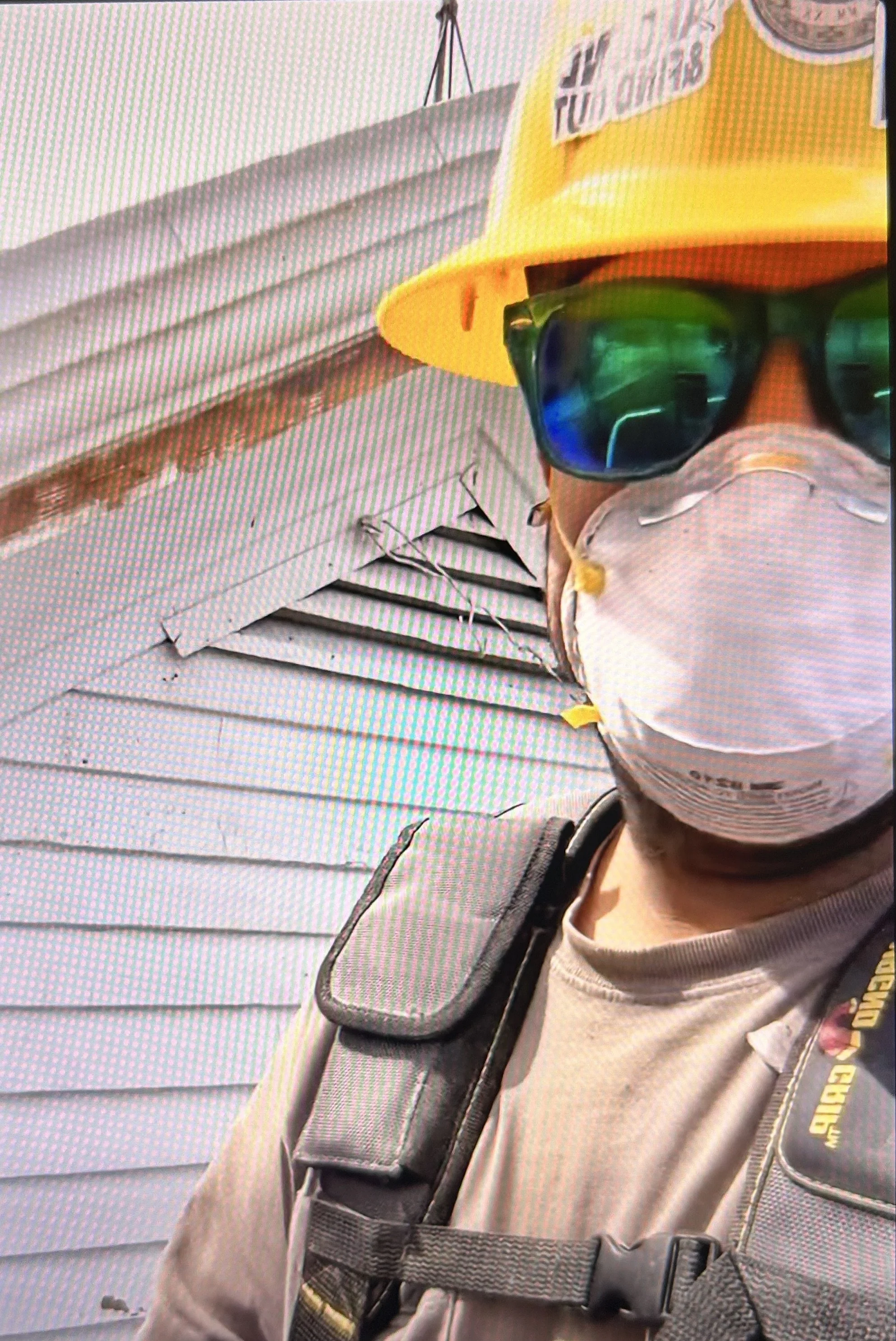 Close-up of a person wearing a yellow hard hat, dark sunglasses, and a gray shirt, with a gray backpack, in front of a building with white horizontal siding.
