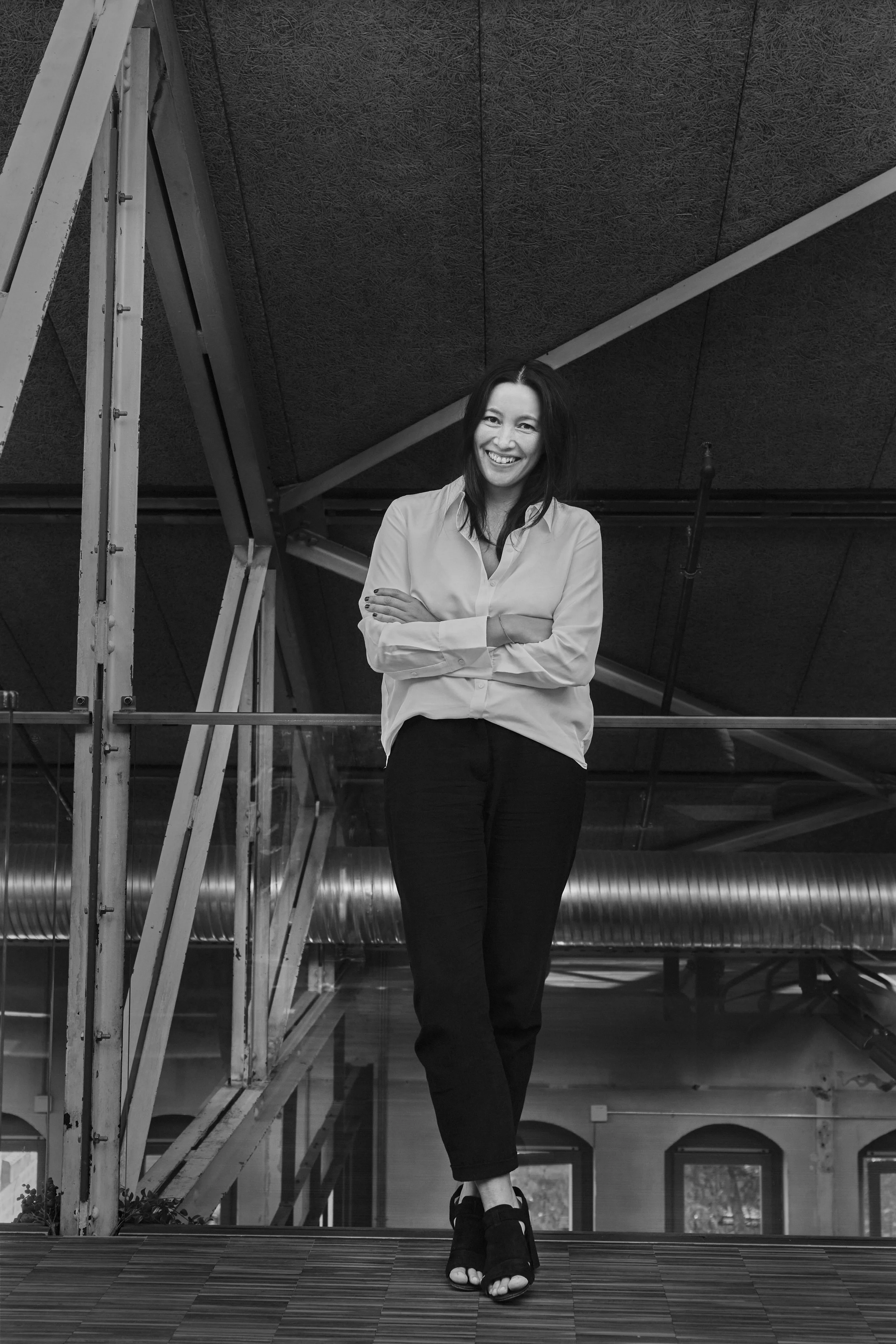 A woman with long dark hair wearing a white blouse and dark pants, standing with crossed arms and smiling, in an industrial-style interior space with metal pipes and a sloped ceiling.