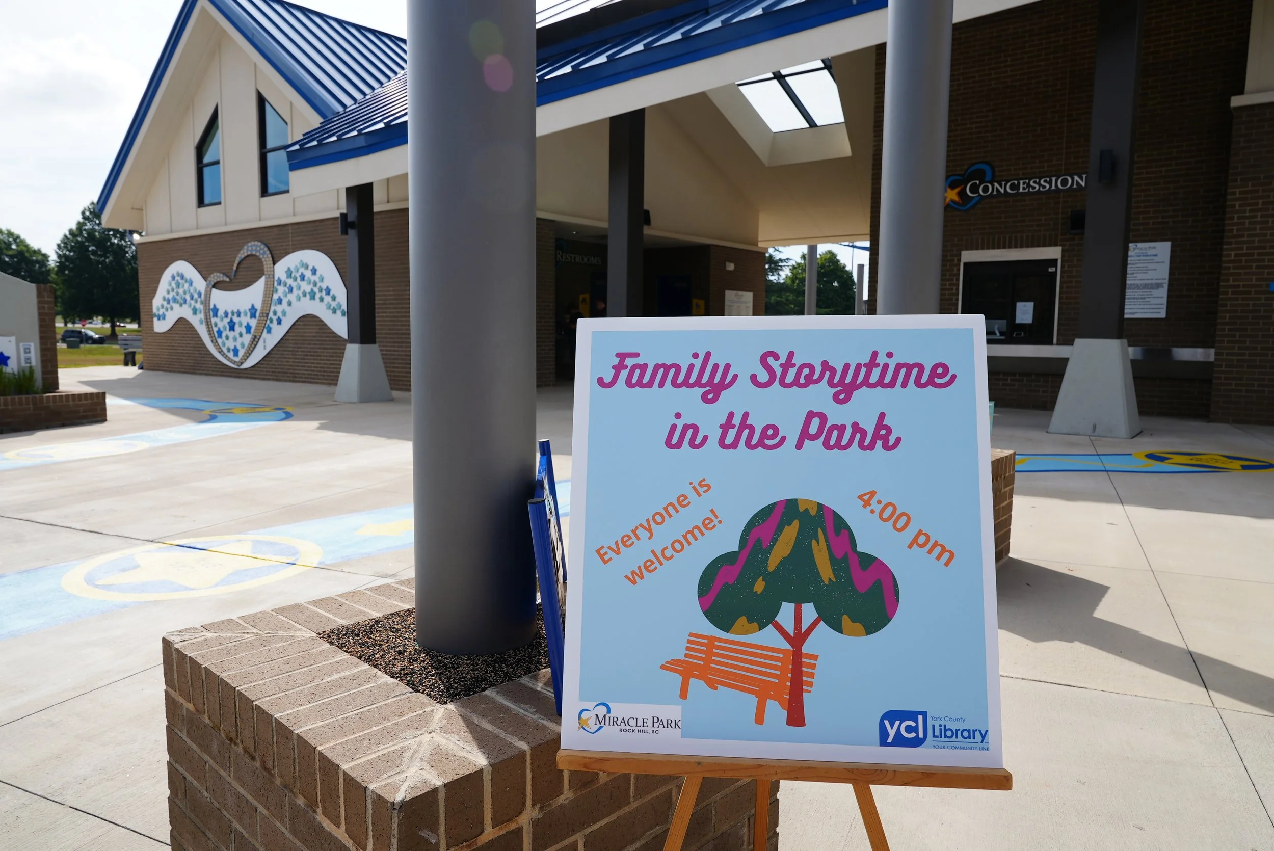 Sign for Family Storytime in the Park outside a building with brick walls and a blue roof, featuring a plaque with a bird and star design and the YCL logo.