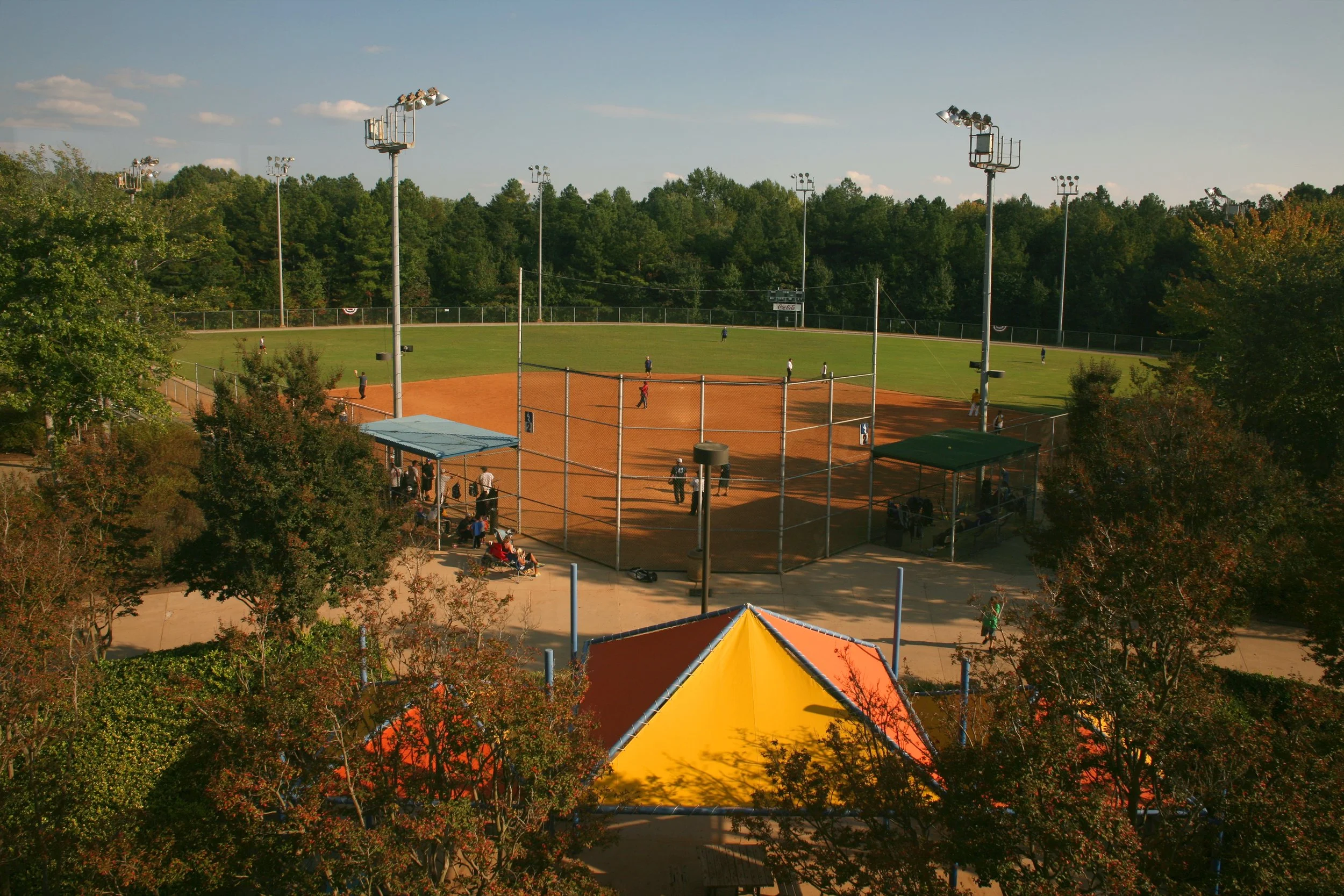 A baseball field with players practicing, surrounded by trees, with large floodlights and a blue sky.