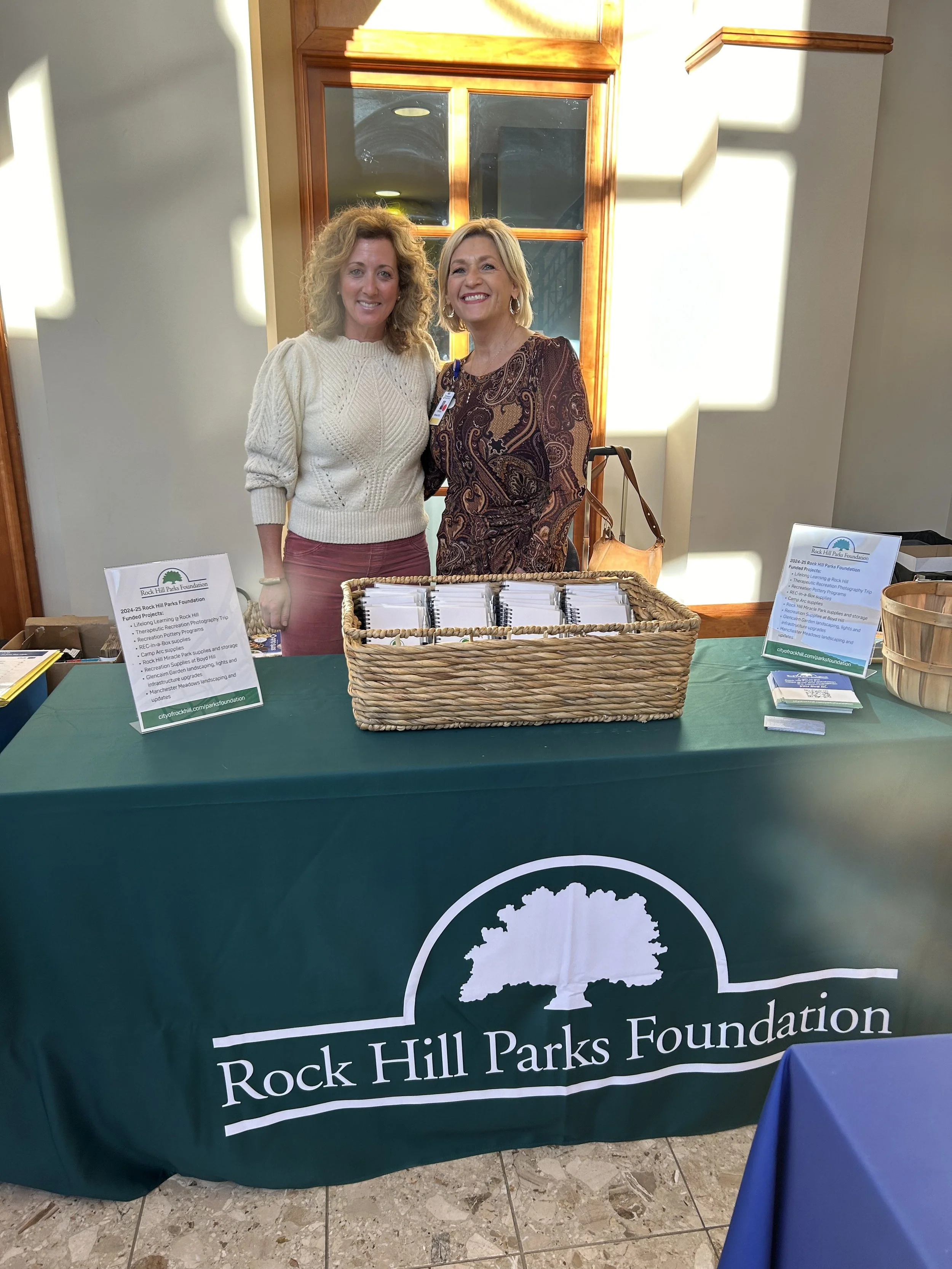 Two women standing behind a table with brochures and signs, representing the Rock Hill Parks Foundation. The table has a dark green tablecloth with a white logo of a tree and the text "Rock Hill Parks Foundation."
