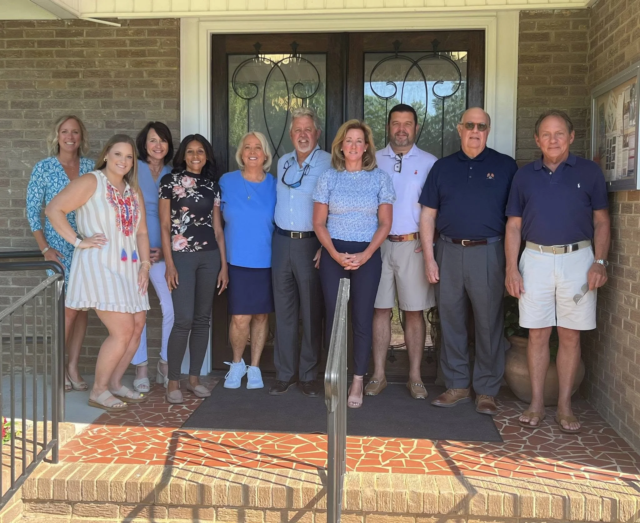 A group of ten smiling people standing on the porch of a brick house in front of a dark wooden door with glass panels. These are some of the members on the Rock Hill Parks Foundation board.