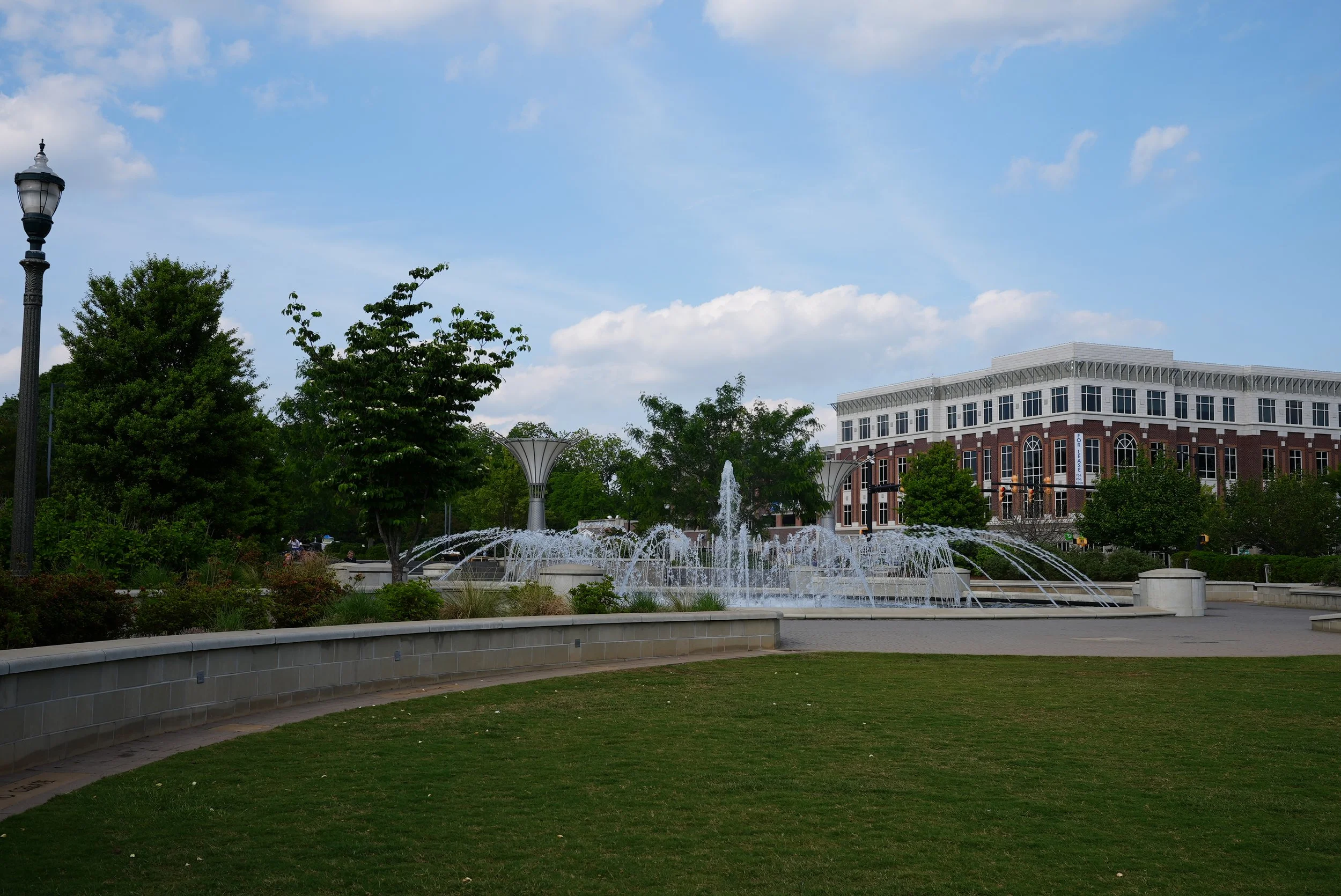 A city park with a fountain in the foreground, surrounded by greenery and trees, with a large modern brick and white building in the background under a partly cloudy sky.