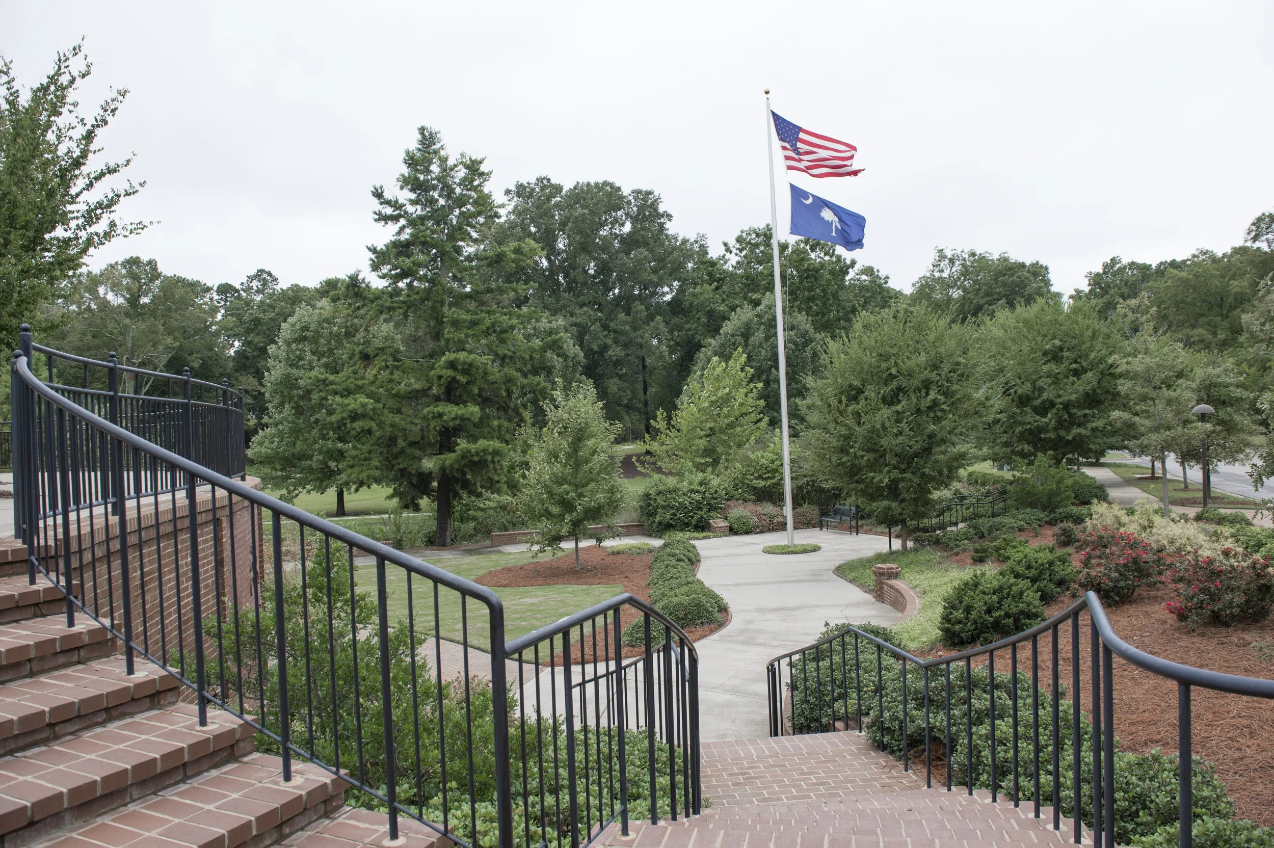 A landscaped park with brick stairs, black metal railings, walking paths, green trees, and two American flags flying on poles against a cloudy sky.