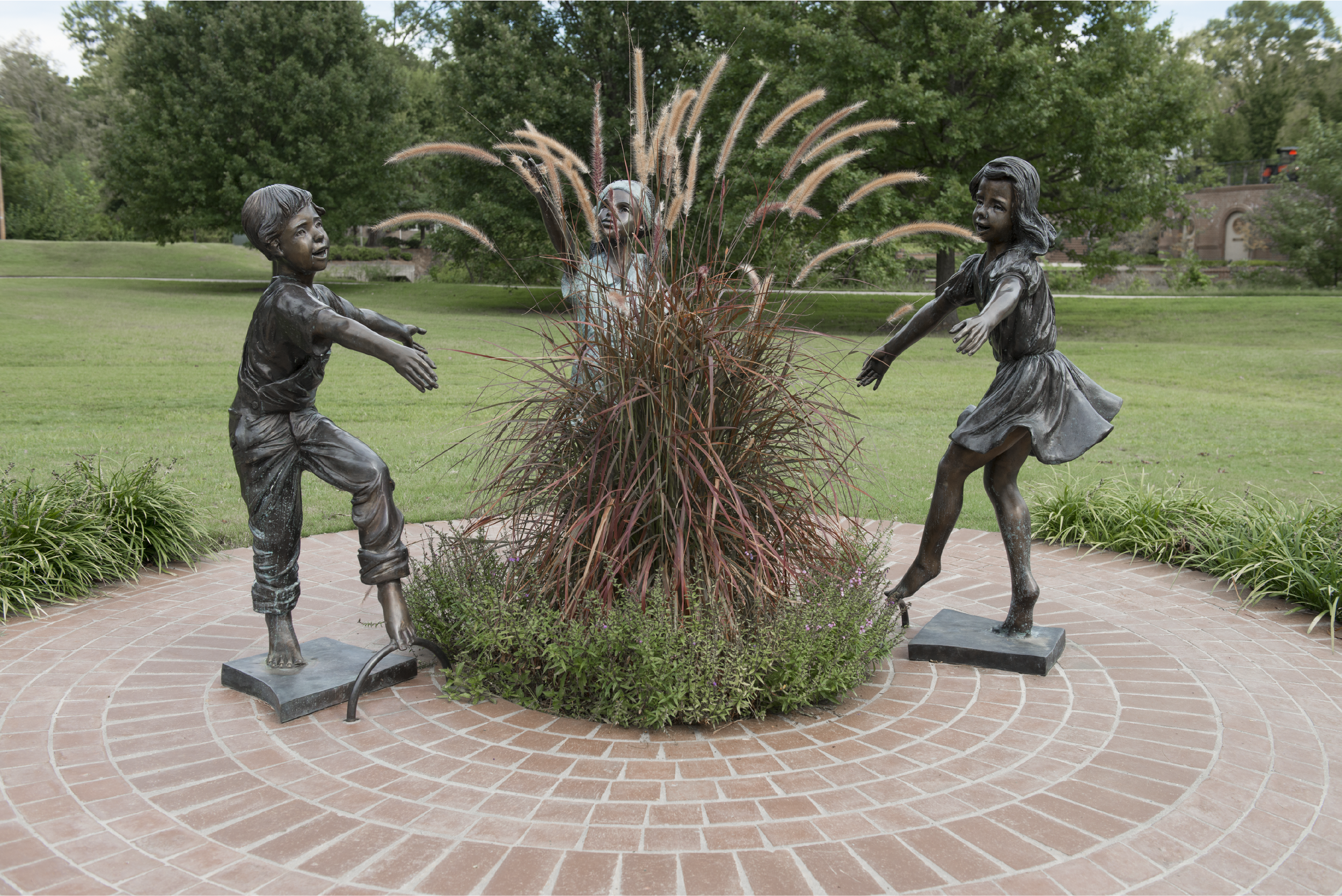 Bronze statues of a boy and girl playing, surrounded by tall ornamental grass, on brick pavement with a grassy park and trees in the background.