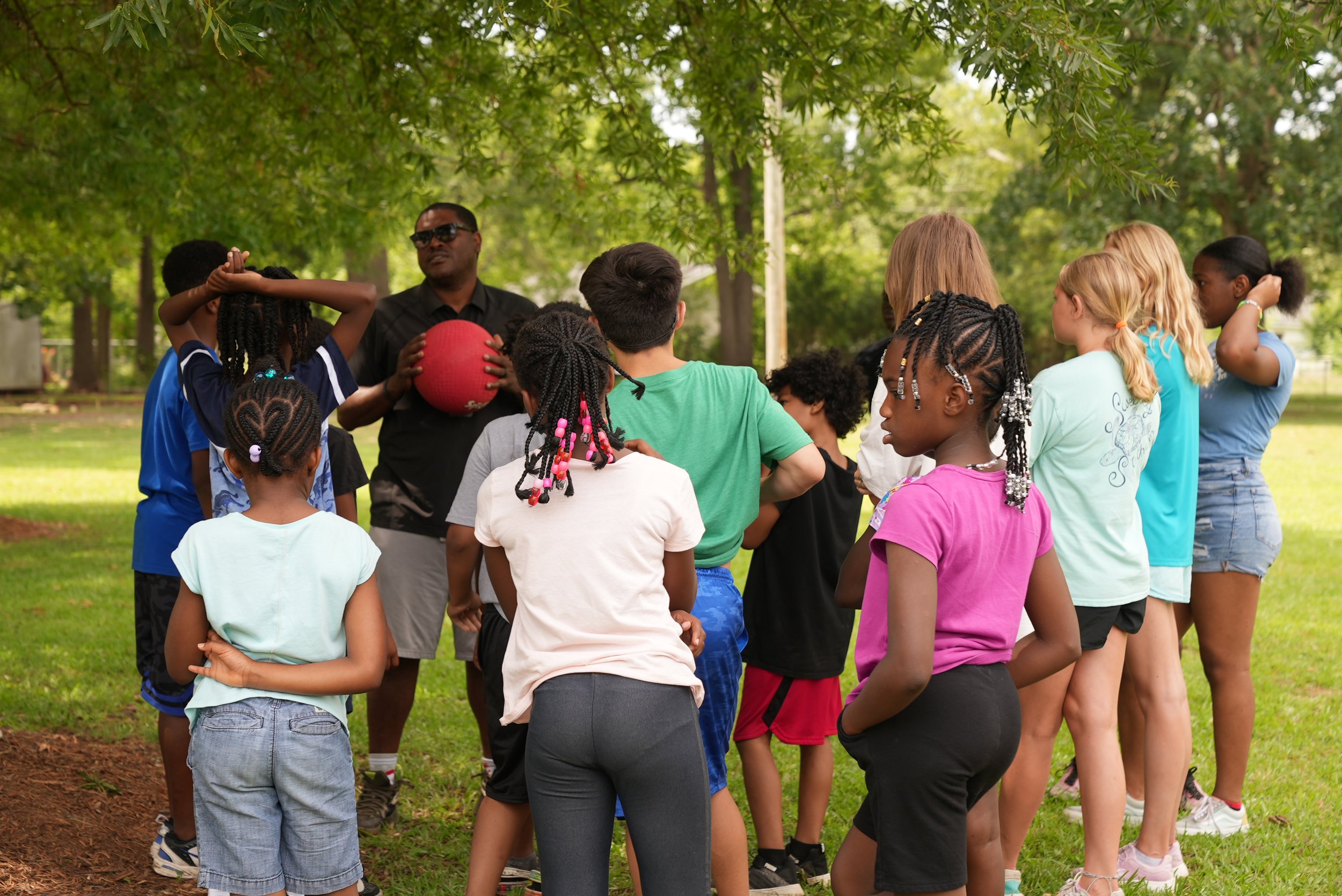 Group of children and an adult in a park, gathered around a man holding a red ball, with green trees overhead.