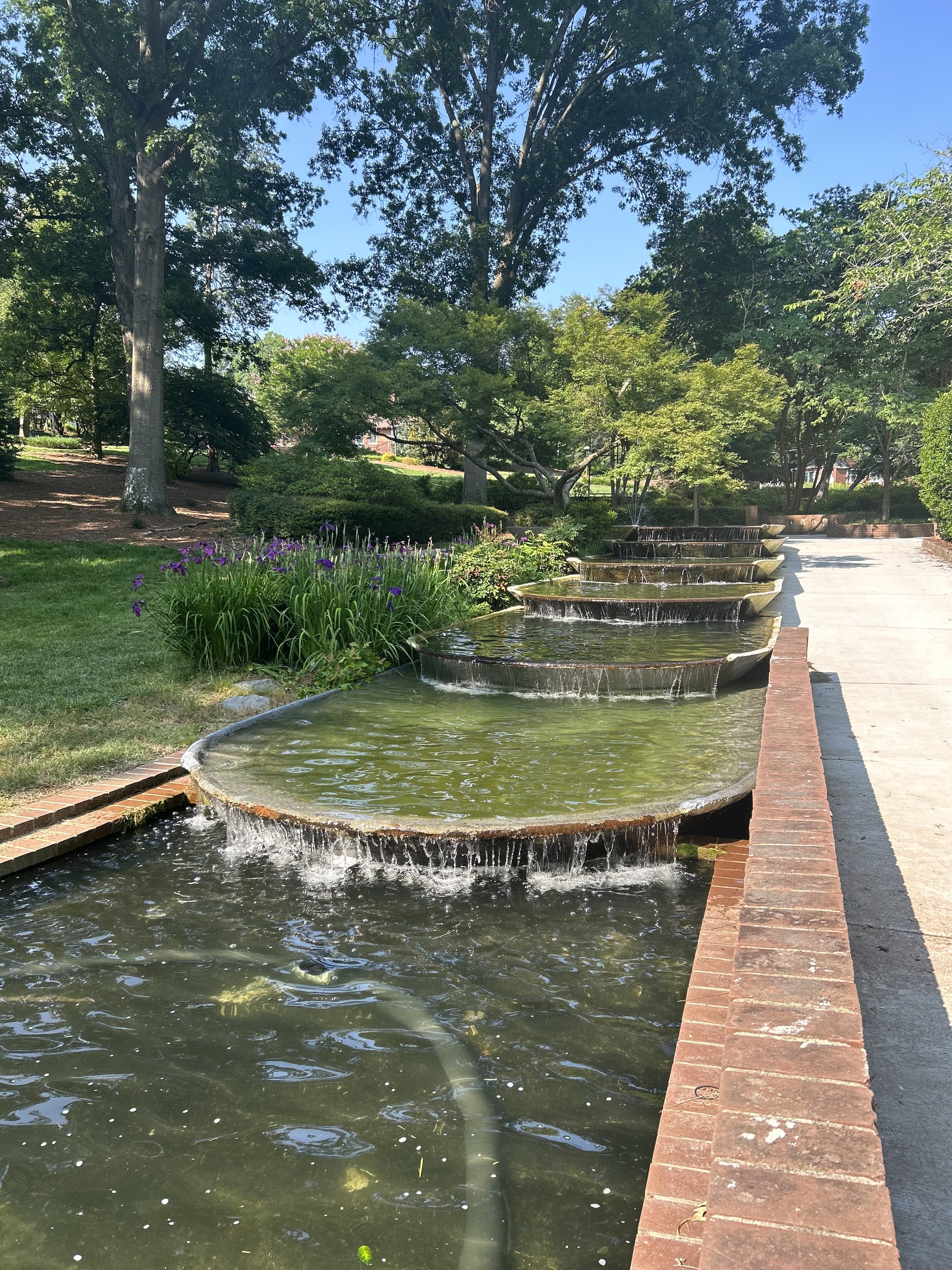A park with a small cascading water feature, surrounded by green grass, purple flowers, and tall trees with dense foliage under a clear blue sky.
