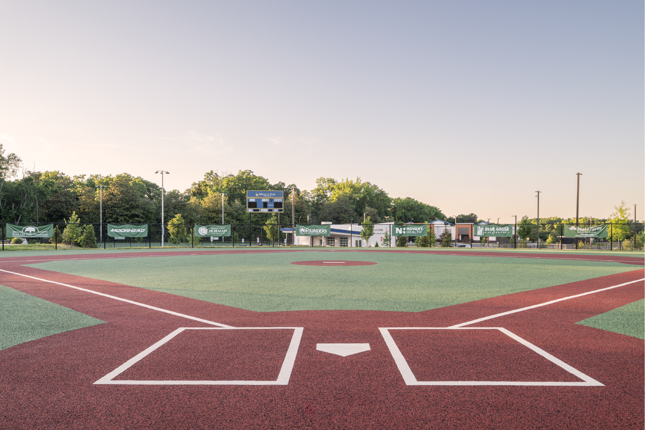 An empty baseball or softball field with a green and red infield, surrounded by green trees and banners, and a scoreboard in the background, during daylight hours.