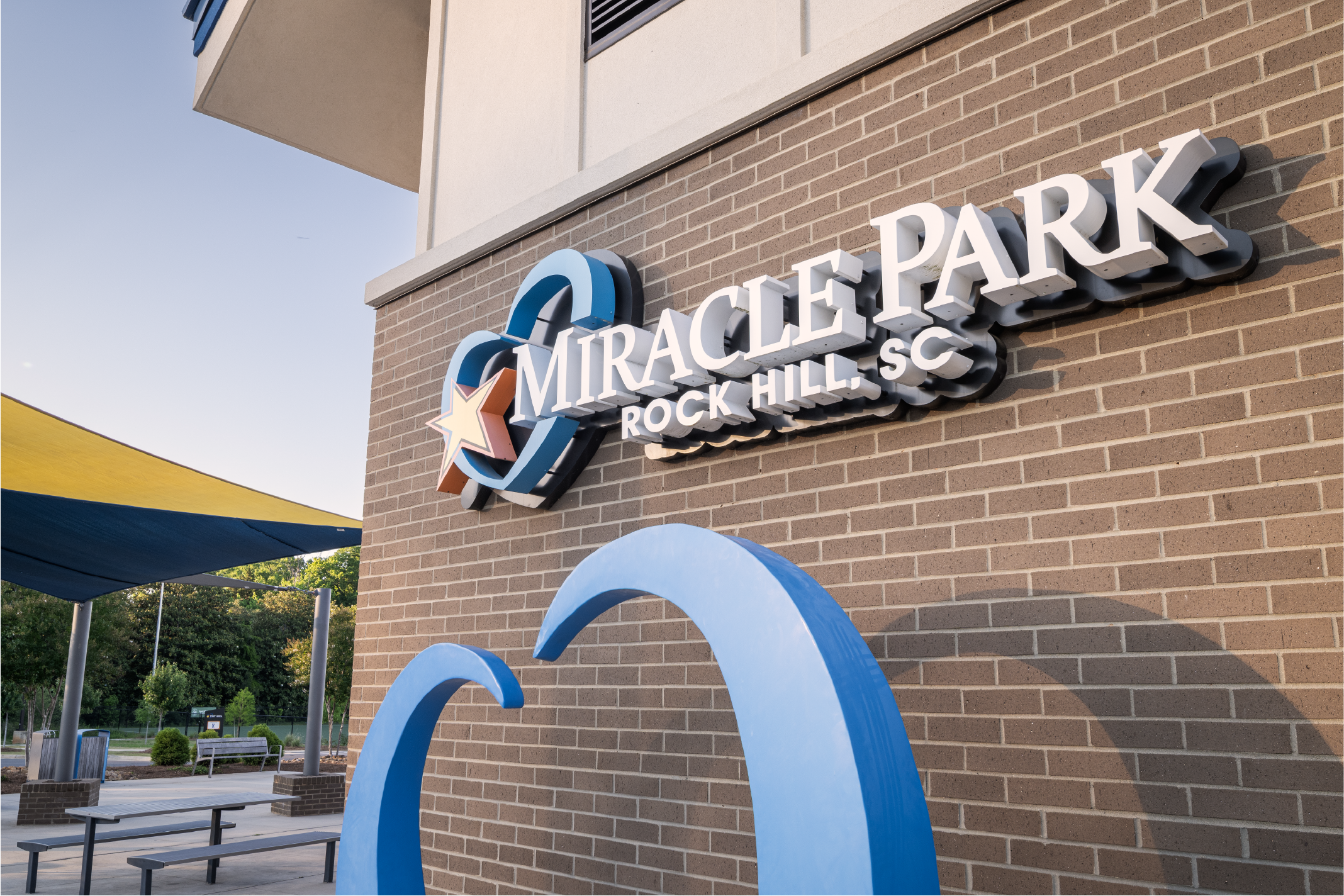 Sign on a brick wall reading 'MIRACLE PARK, ROCK HILL, SC' with decorative blue arches nearby, outdoor seating and trees in the background.