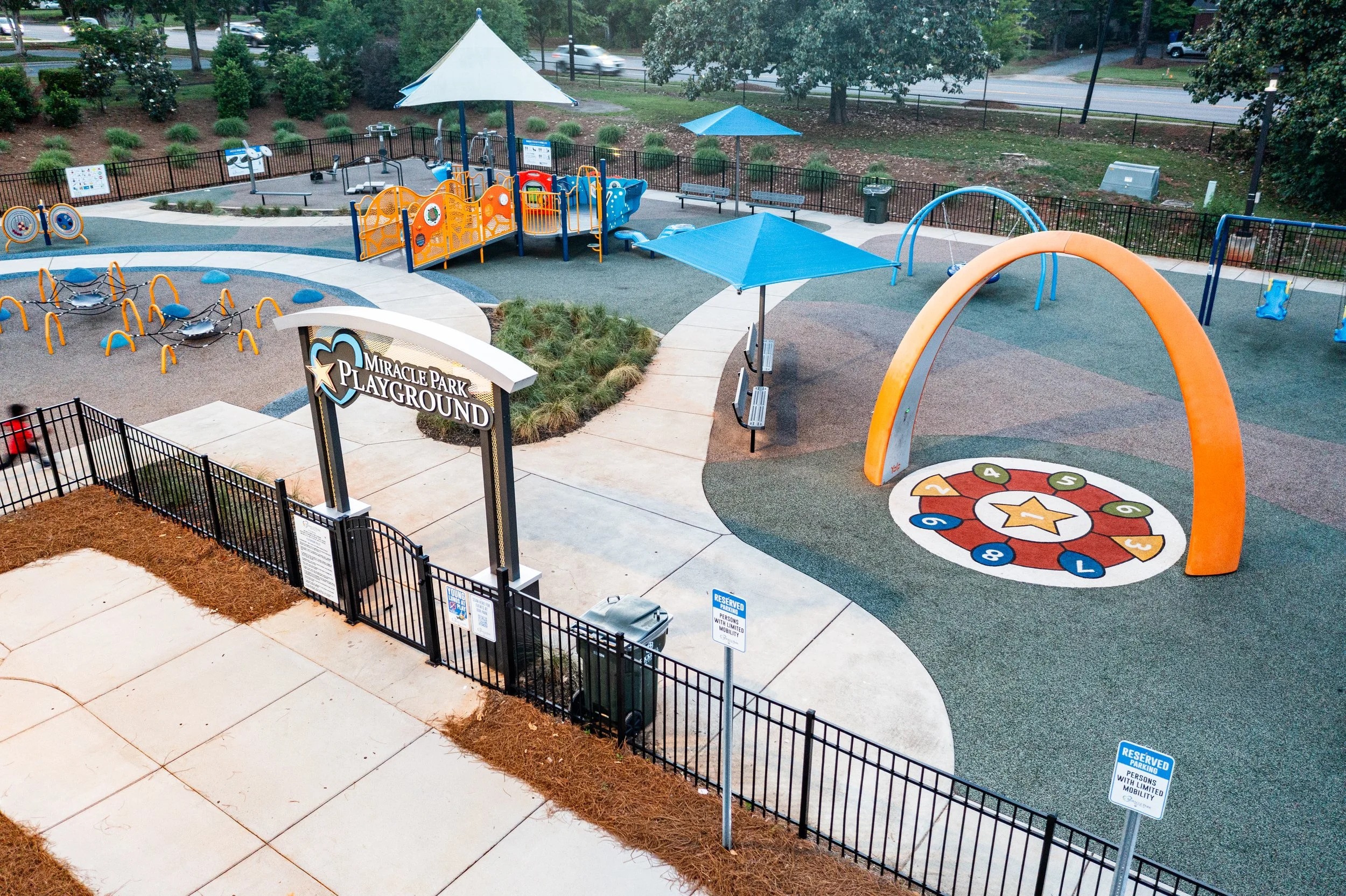 Children's playground with climbing structures, slides, a large orange and blue arch, a circular game area with numbered sections, benches, and shaded areas. Sign that reads 'Miracle Park Playground' at the entrance, fenced enclosure, and trees in the background.
