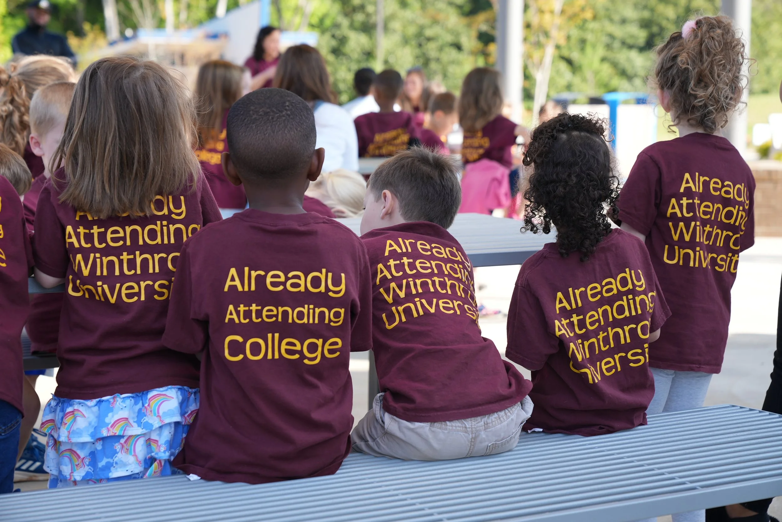 Group of children sitting on a silver bench outdoors, wearing maroon shirts that read 'Already Attending Winthrop University,' with trees and a person speaking at a podium in the background.