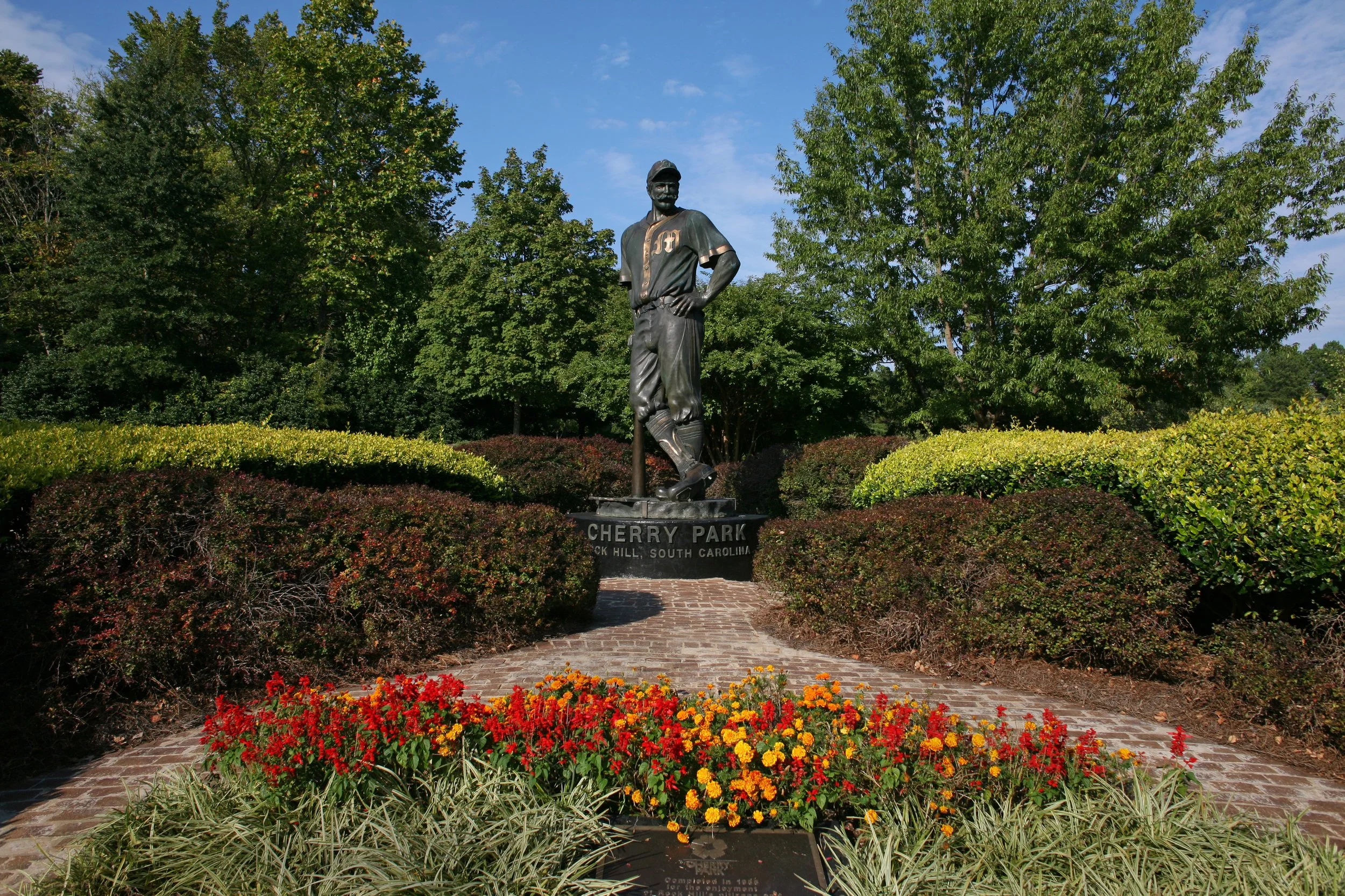 Statue of a baseball player at Cherry Park in Rock Hill, South Carolina, surrounded by colorful flowers, trees, and a clear blue sky.