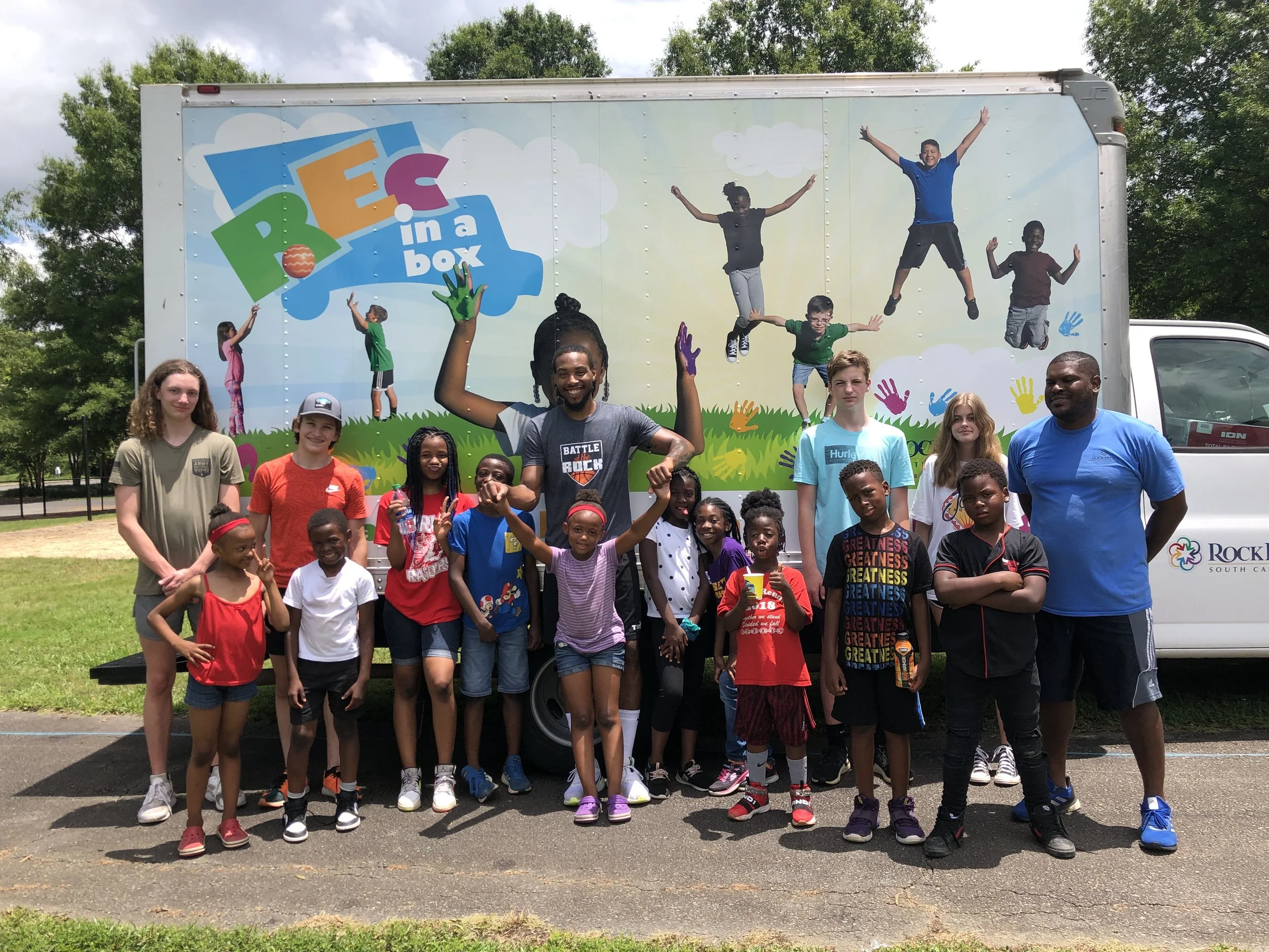 Group of children and adults posing in front of a mobile activity truck with a colorful sign that reads 'REC in a box'.