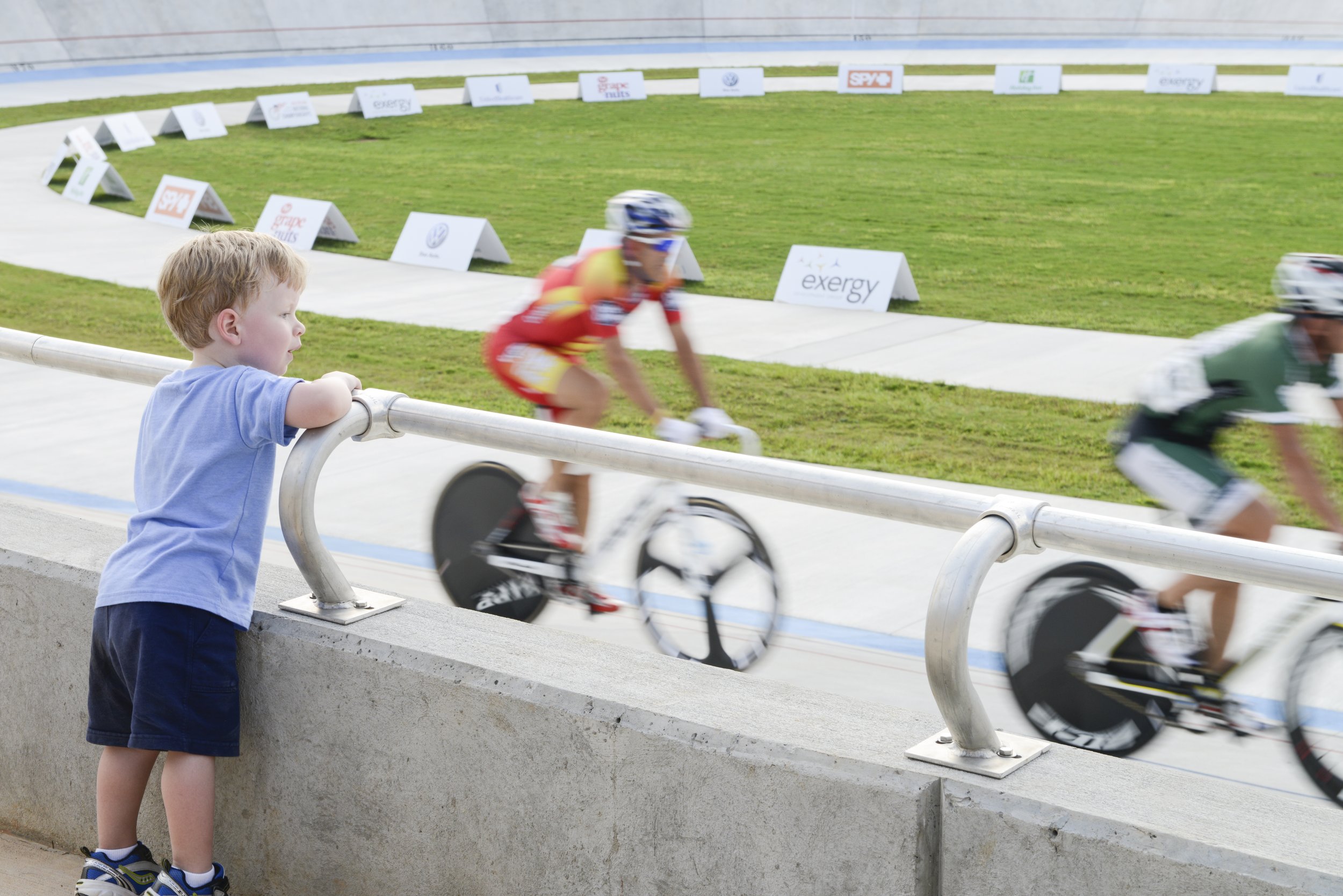 A young boy in a blue shirt and shorts watching cyclists race on a velodrome track, leaning on a metal railing.
