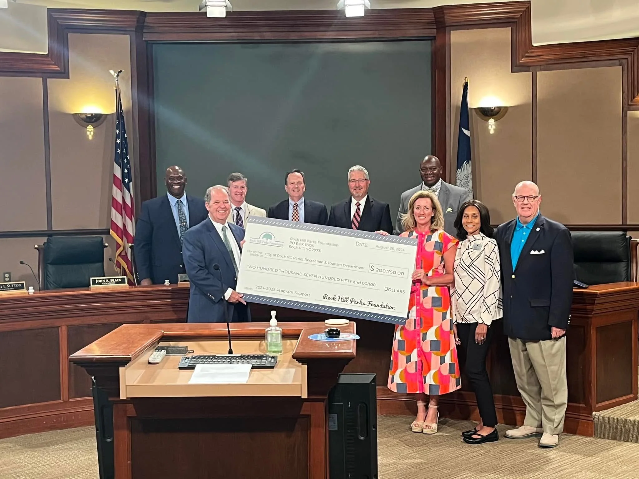 A group of nine people standing in a conference room, holding a large check made out to the City of Rock Hill Parks, Recreation & Tourism Department. The room has wood-paneled walls, a large green chalkboard, American flags, and a desk with microphones and nameplates.
