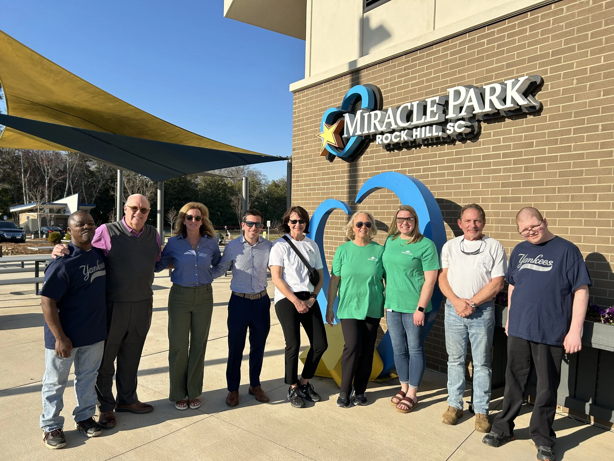 A group of nine people standing in front of a brick wall with a sign that reads 'Miracle Park Rock Hill SC'. The group is outdoors on a sunny day, with colorful shade sails overhead.