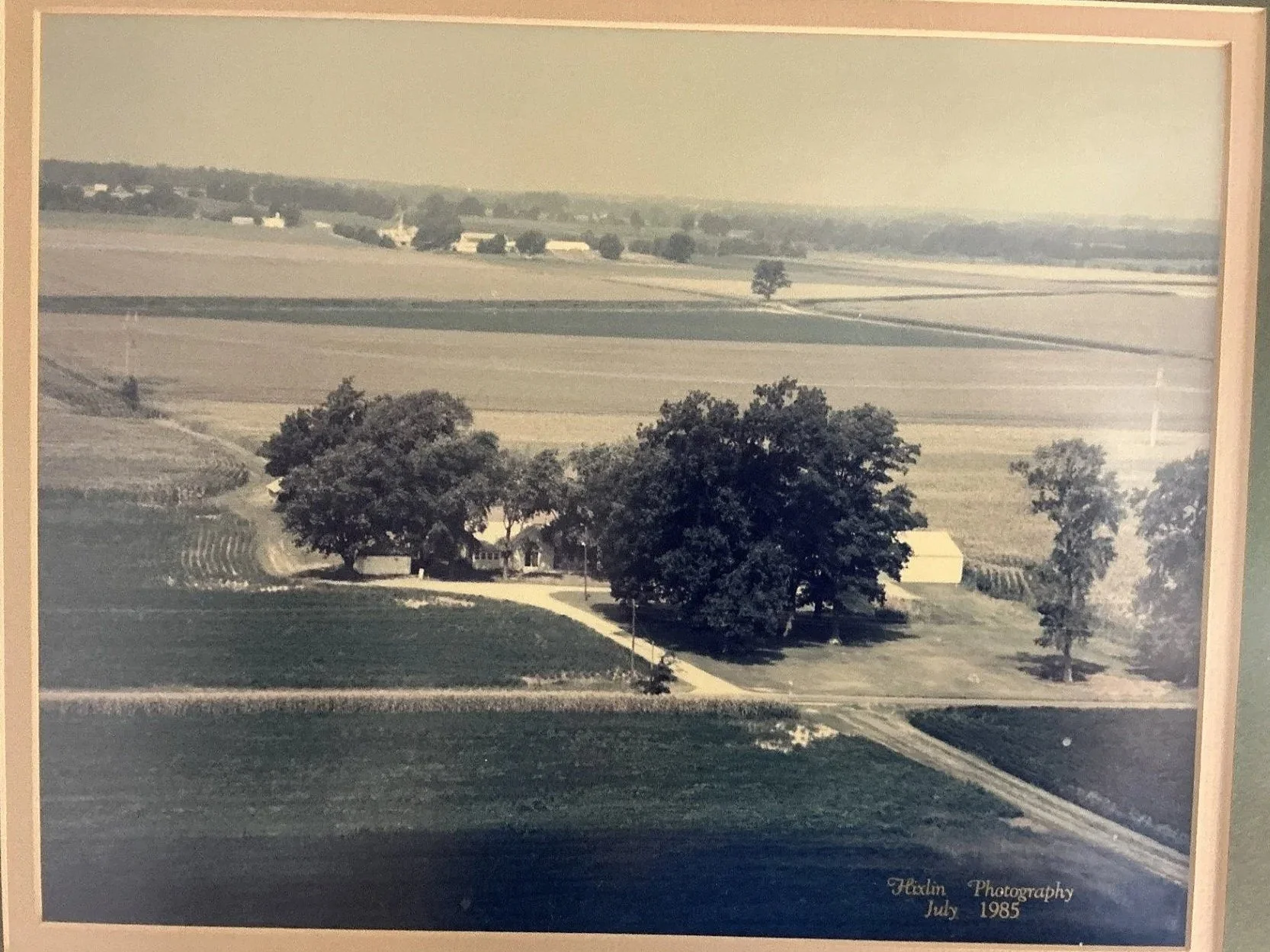 A black and white photograph of a rural landscape, showing rolling fields, farm buildings, and a few trees. The photo has text at the bottom right corner that reads "Hexton Photography July 1985."