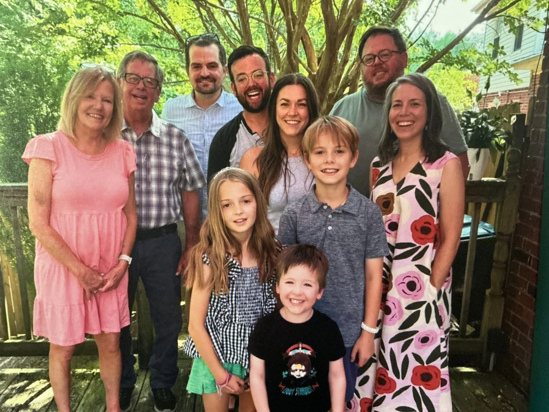 A group of nine people, including women, men, and children, posing outdoors in front of a green leafy tree and a wooden fence, smiling at the camera.