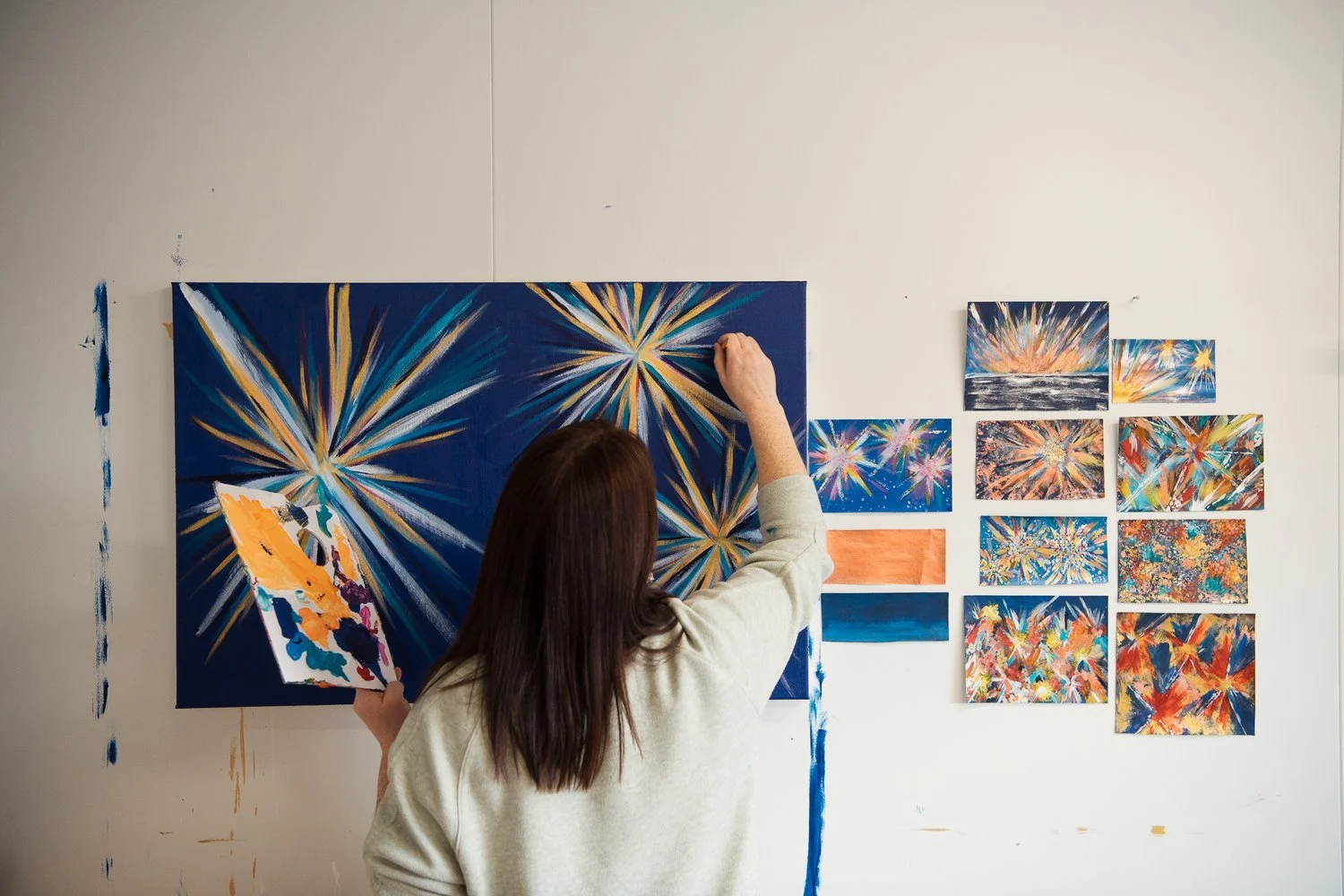 An artist paints fireworks on a large canvas in a studio, with smaller painted canvases of fireworks arranged on the wall nearby.