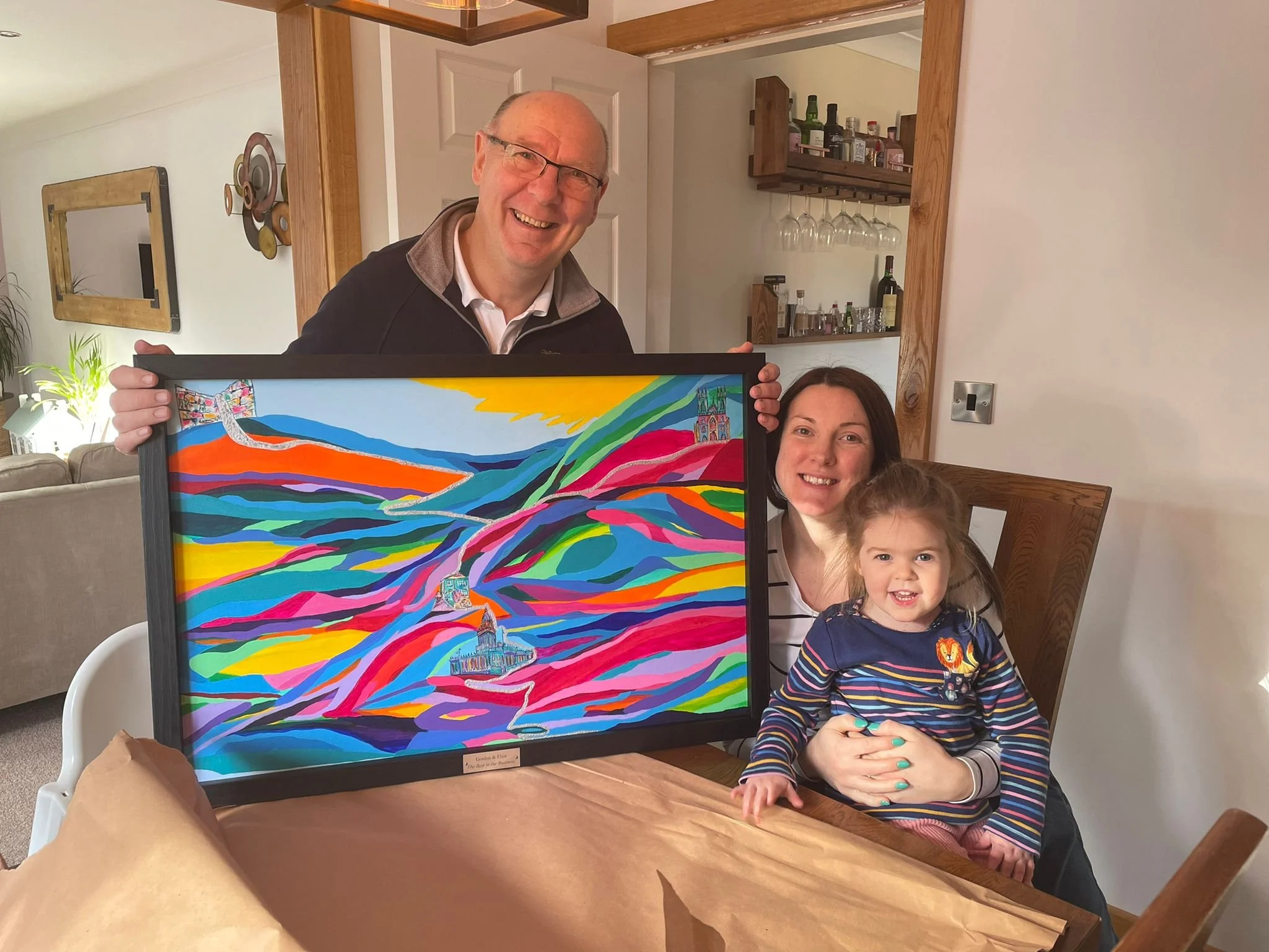 A smiling older man and a young girl are posing together inside a home, holding a colorful framed artwork. The artwork features a vibrant landscape scene with rolling hills, a cathedral, and abstract colorful patterns. The man is wearing glasses and a dark jacket, while the girl is smiling and dressed in a striped top with a lion embroidery.
