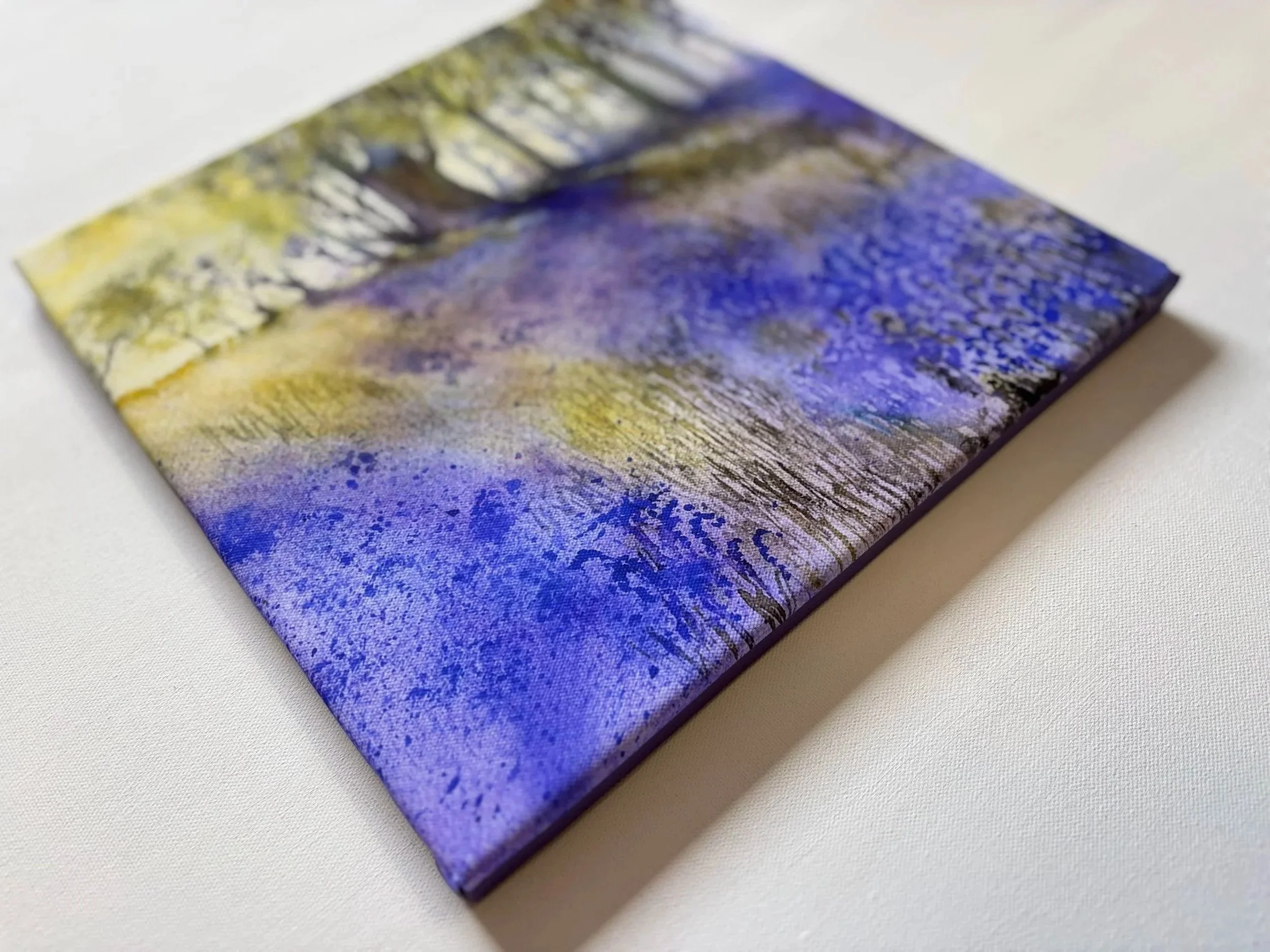 Close-up of a small, square canvas print with a blurry, abstract landscape scene in yellow, green, purple, and blue colors resting on a white surface.